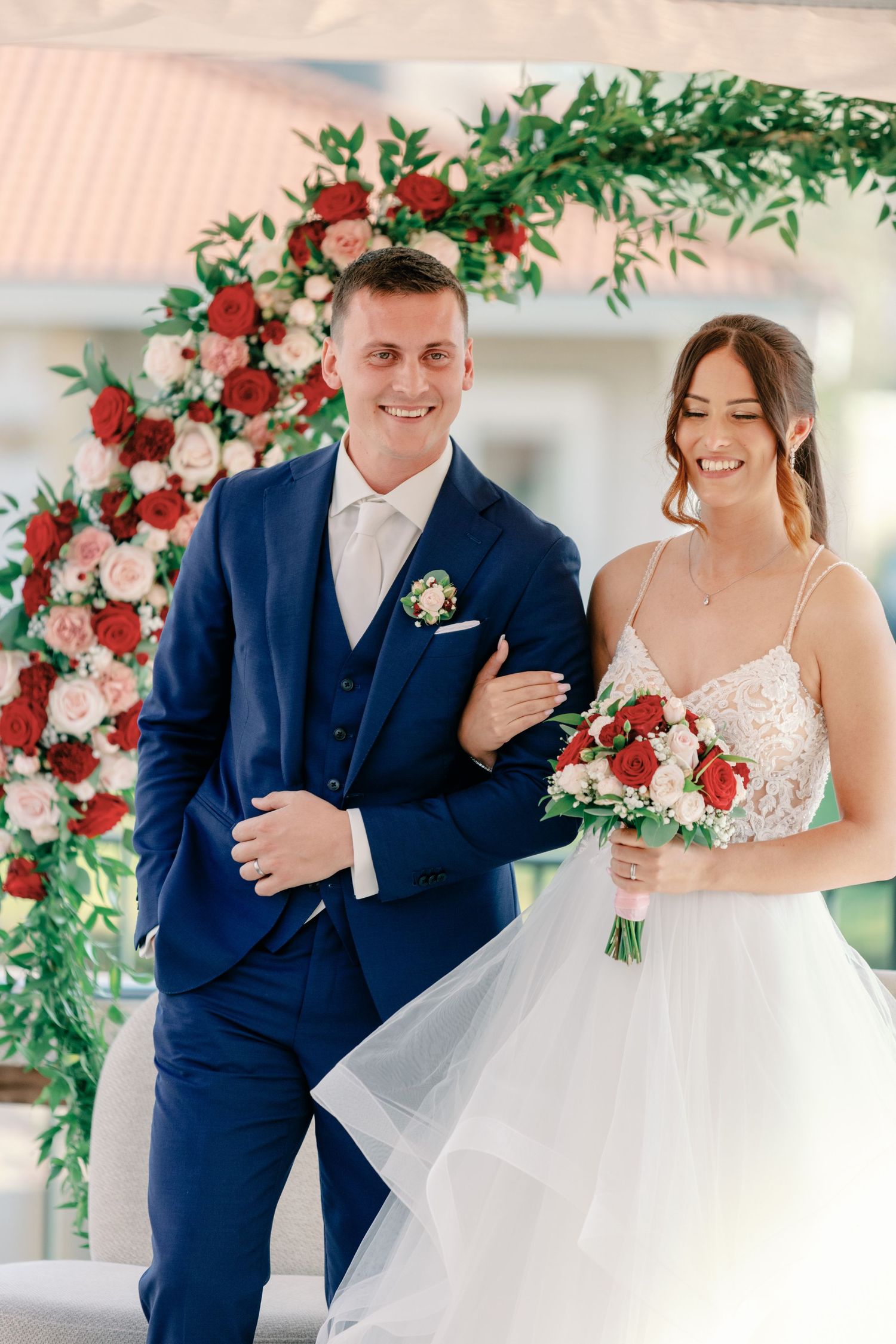 Newlyweds celebrate under floral arch with red roses and greenery at outdoor venue.