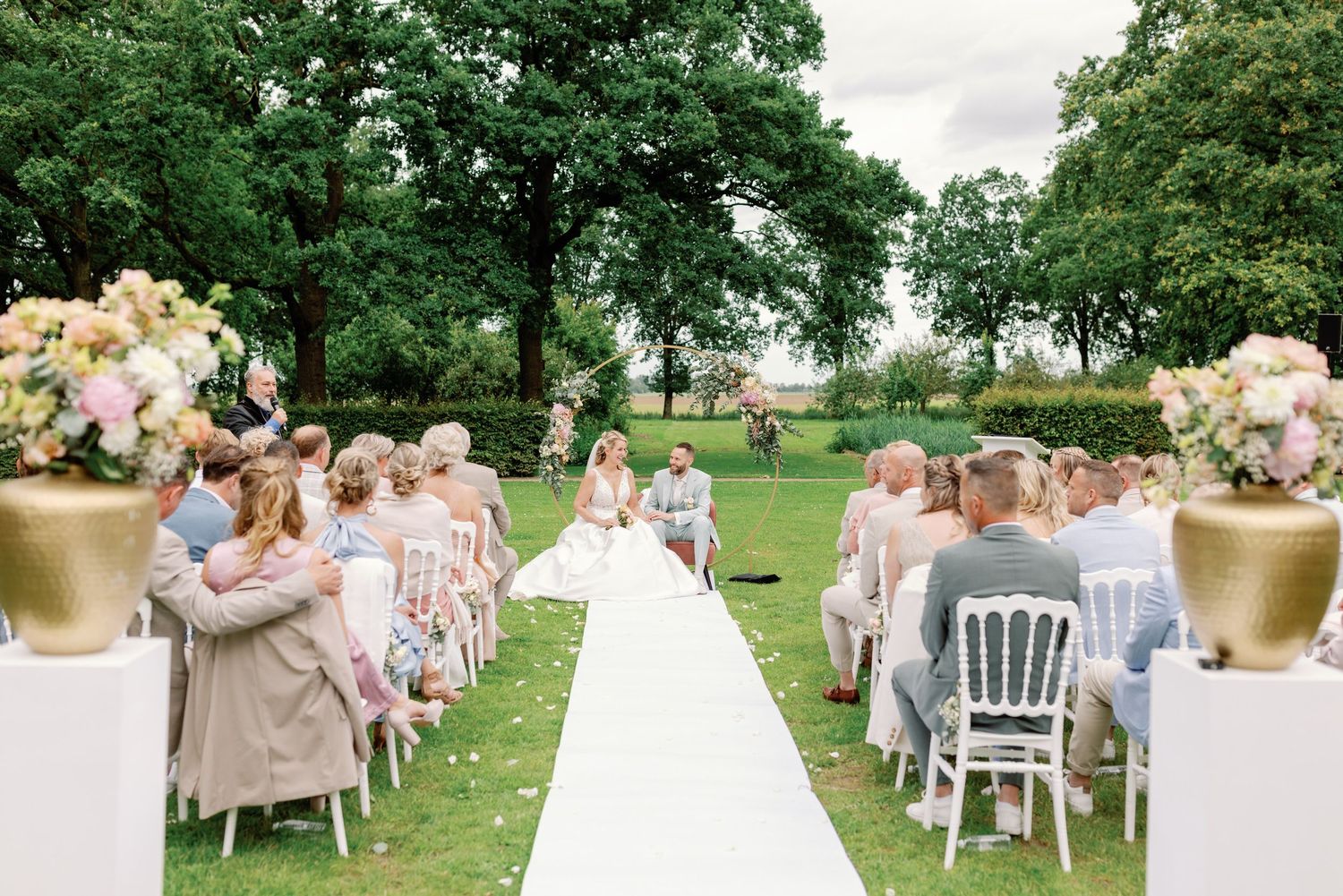 Outdoor garden wedding ceremony setup with white aisle runner, gold floral arrangements, and guests seated on white chairs.