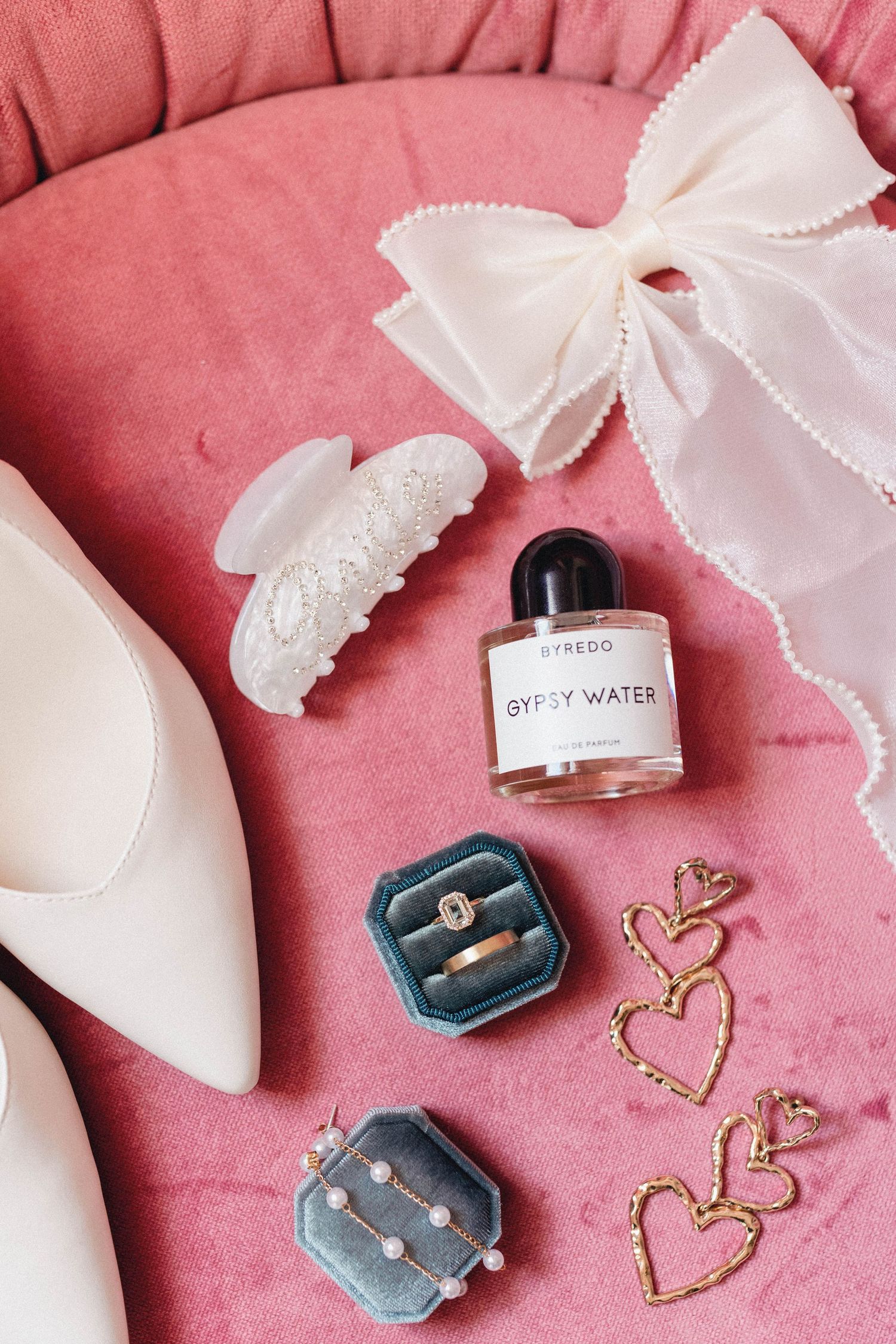 Perfume bottle and gold heart decorations arranged on pink textured background with white ribbon accents.