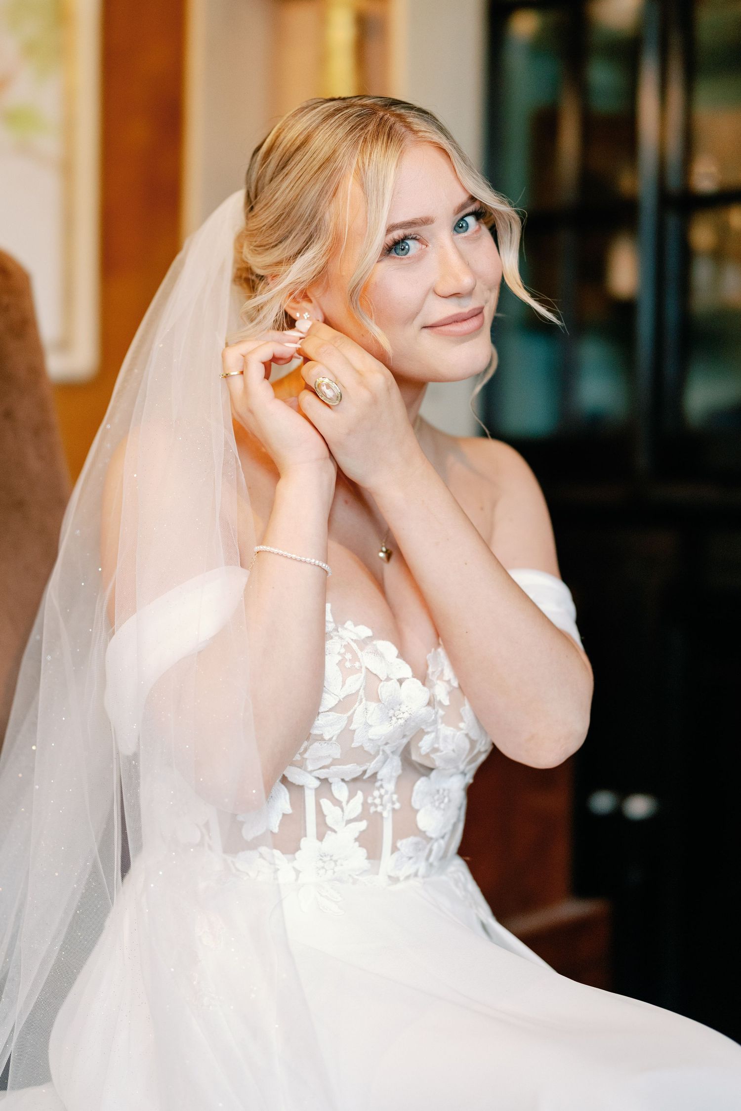 A bride in a white lace wedding dress with sheer veil adjusts her earrings while getting ready.