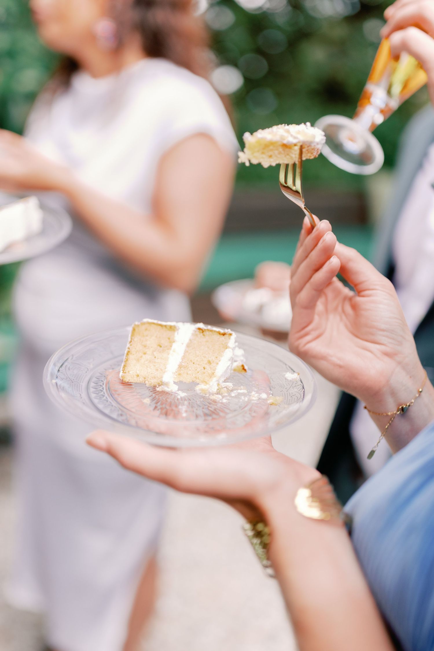 A hand holds a delicate plate with a slice of vanilla wedding cake while another hand lifts a bite with a fork.