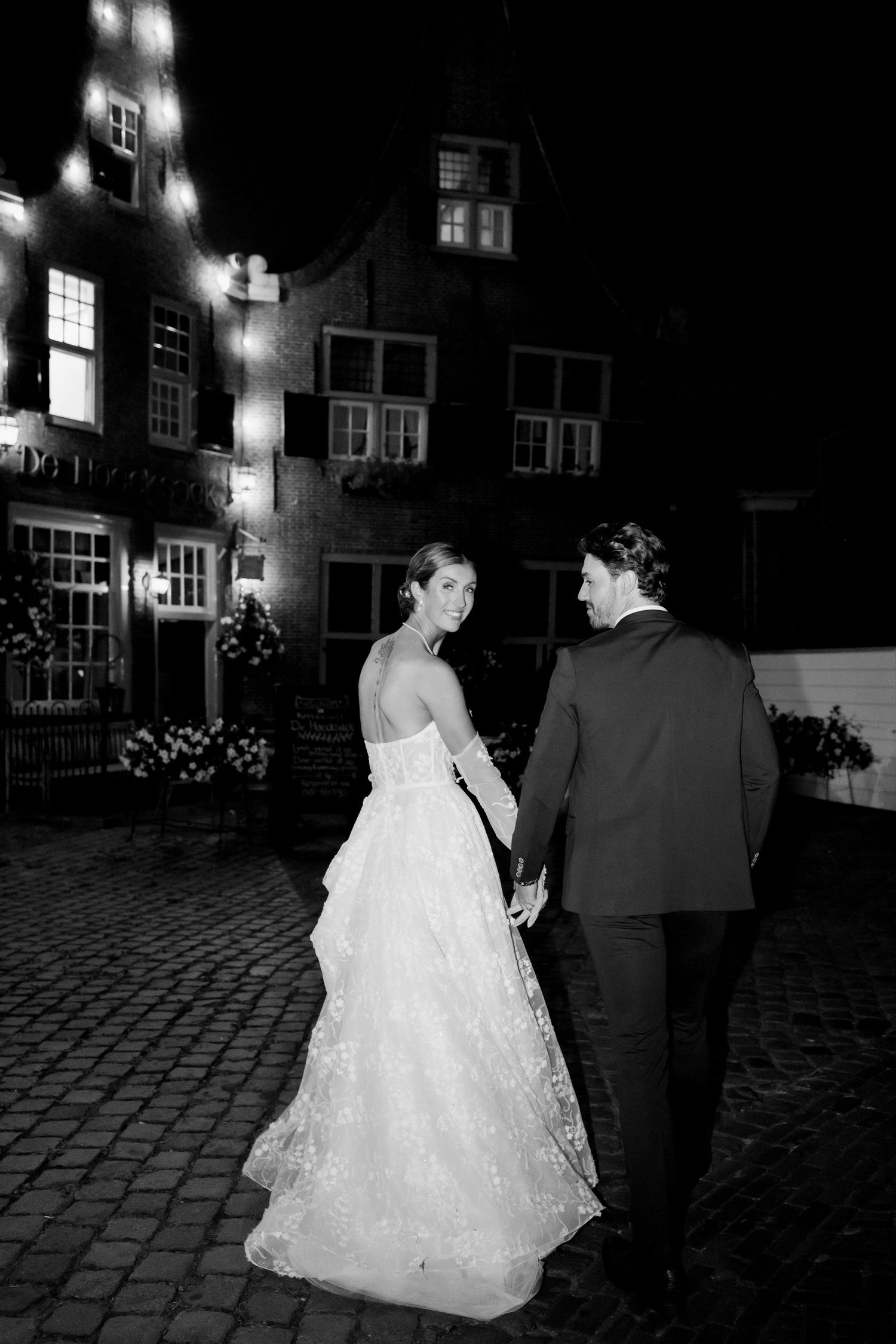 A bride in a white ballgown and groom in dark suit walk together on a cobblestone street at night.