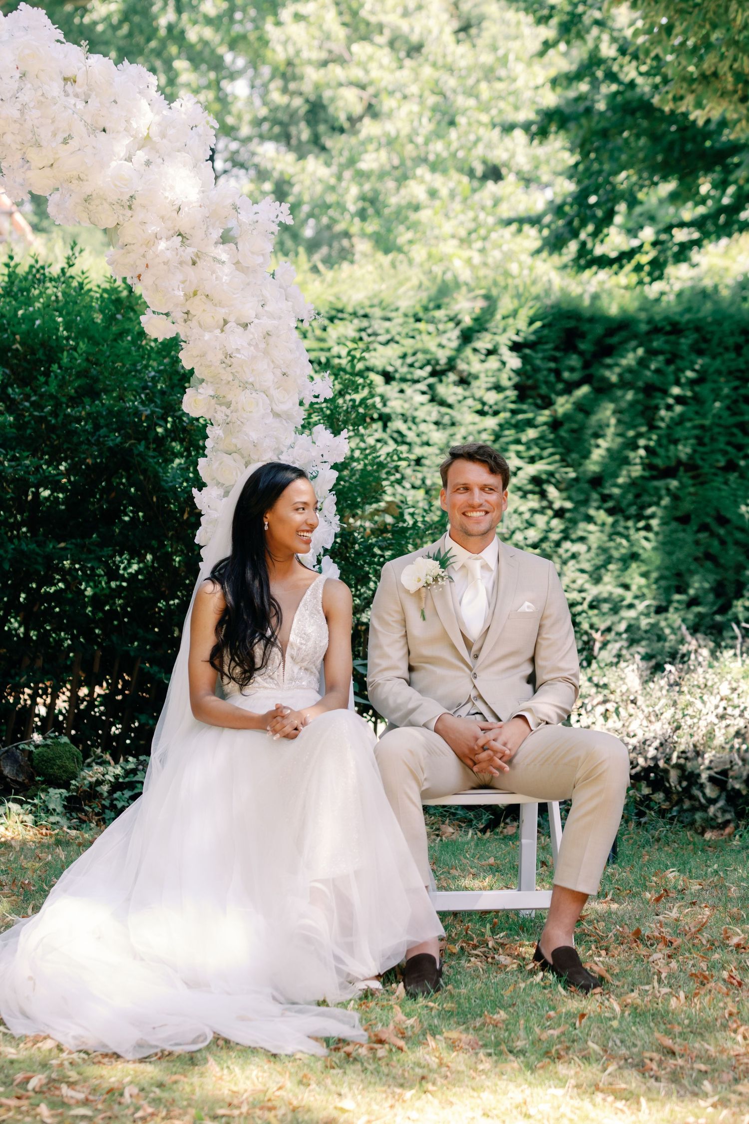 A couple in wedding attire share joyful moments while sitting together in a lush garden setting.