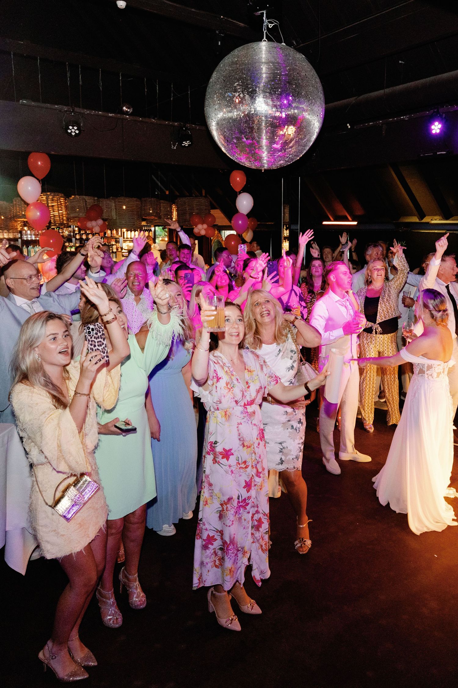 A lively wedding reception dance floor under a disco ball with guests dancing in colorful attire and pink lighting.