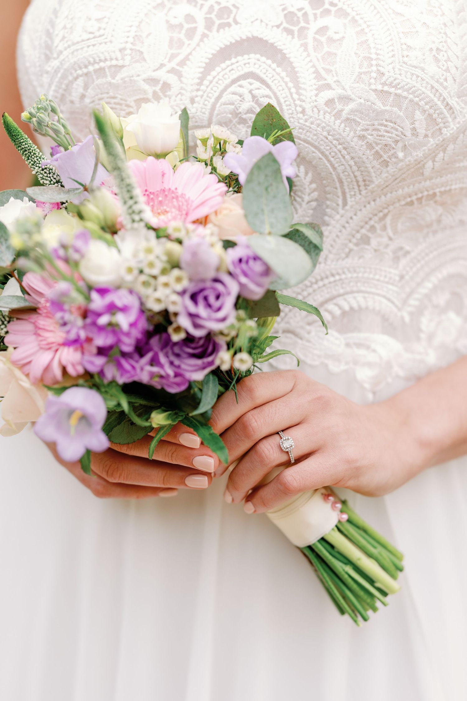 Close up of bridal bouquet with pink and purple flowers against white lace dress with engagement ring visible.