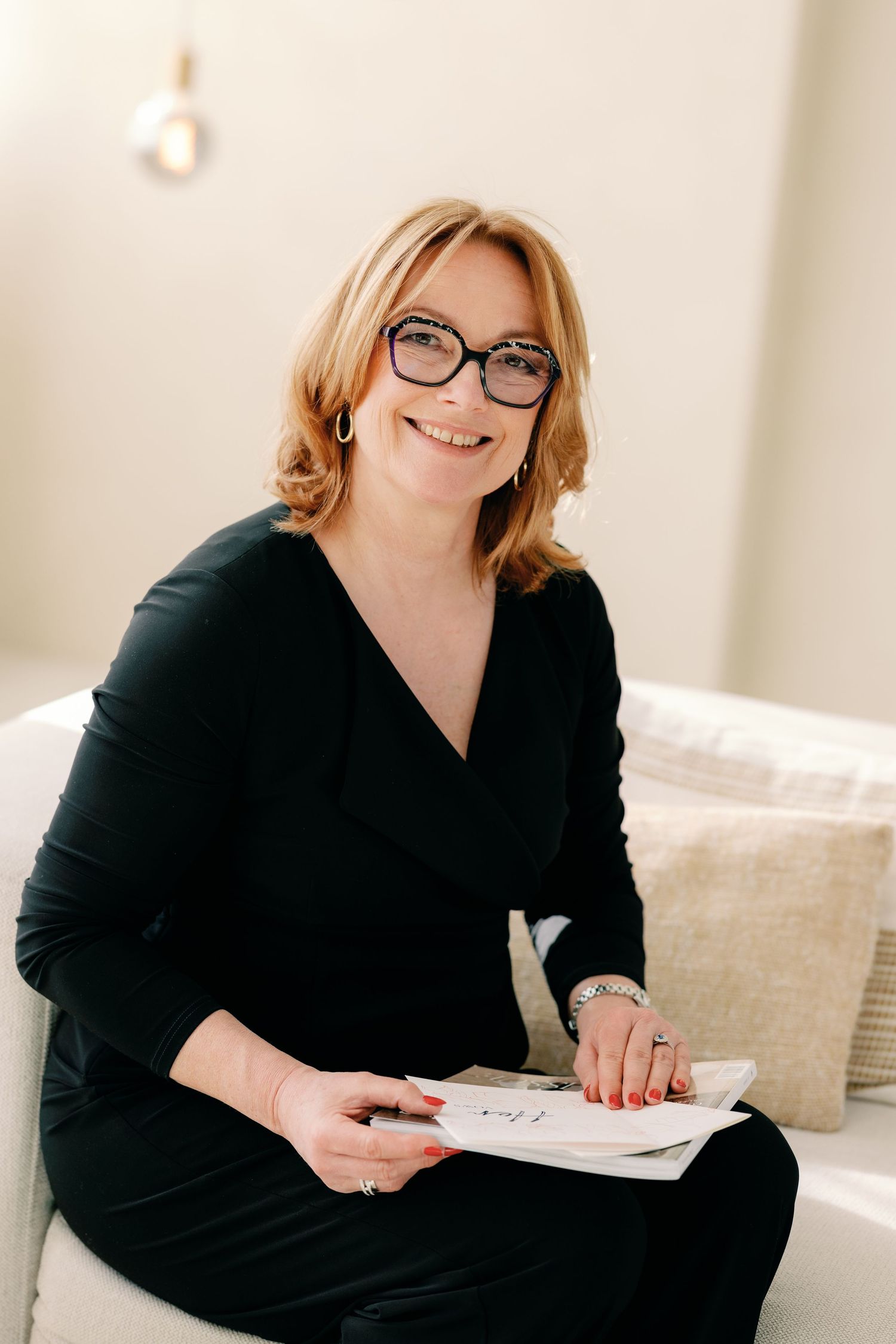Professional wearing glasses and black dress sits on light colored couch with notebook and pen.