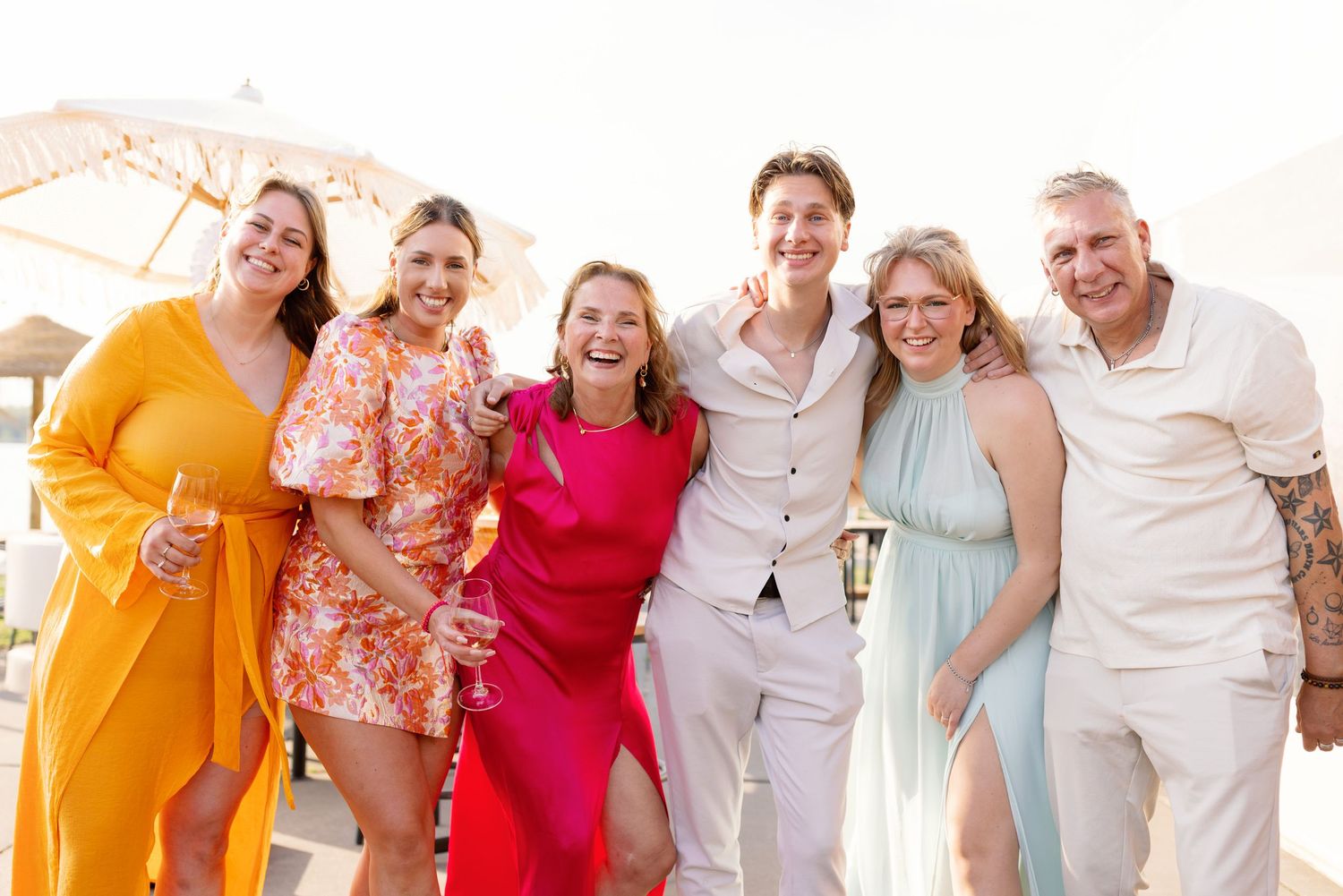 Group of six friends posing together at a sunny beach celebration, wearing colorful outfits in orange, pink, and white.