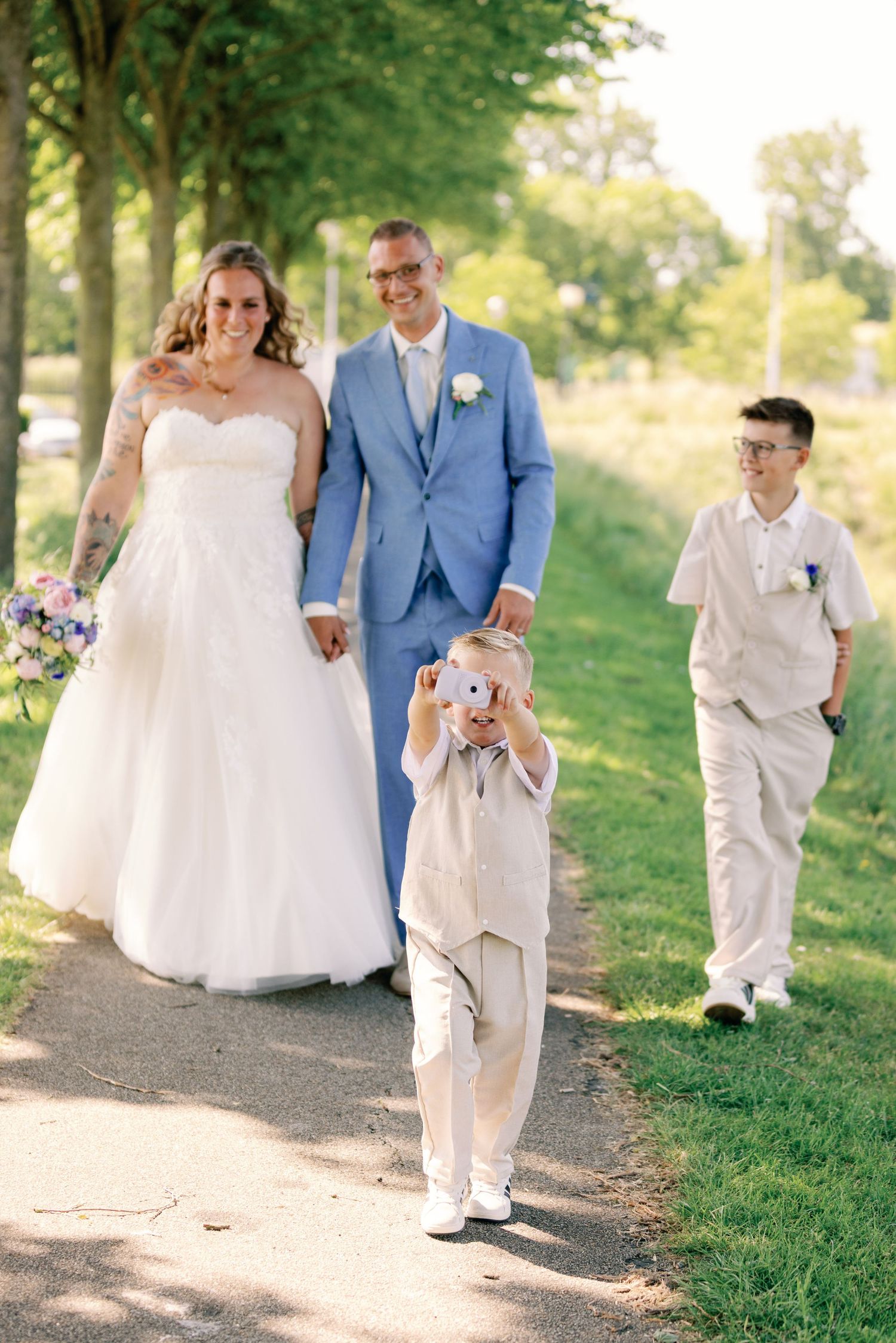 Wedding couple walks with two boys on a tree-lined path, young boy photographing with a pink camera.