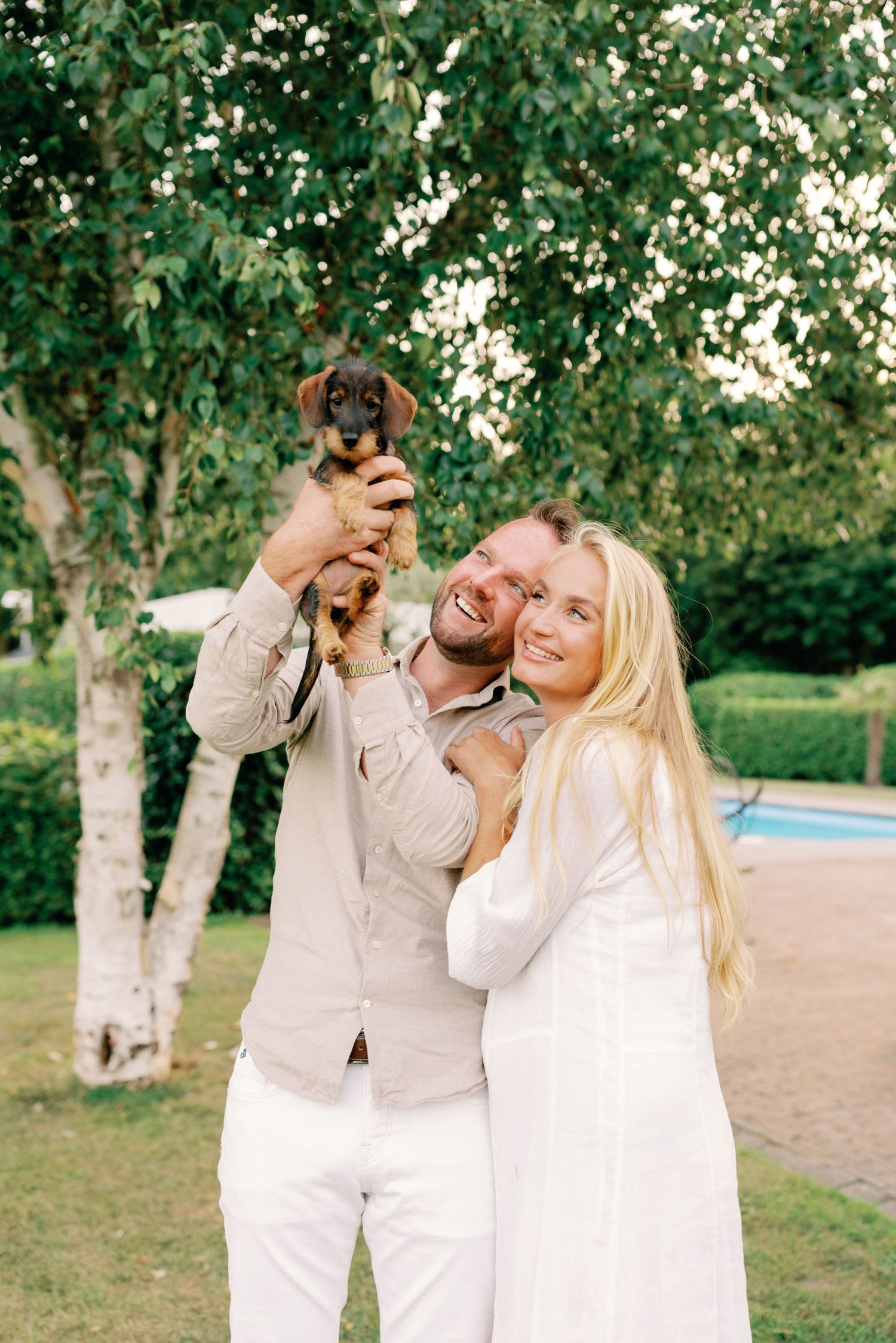 Happy couple holding a dachshund puppy outdoors near birch trees, woman in white dress embracing smiling man.