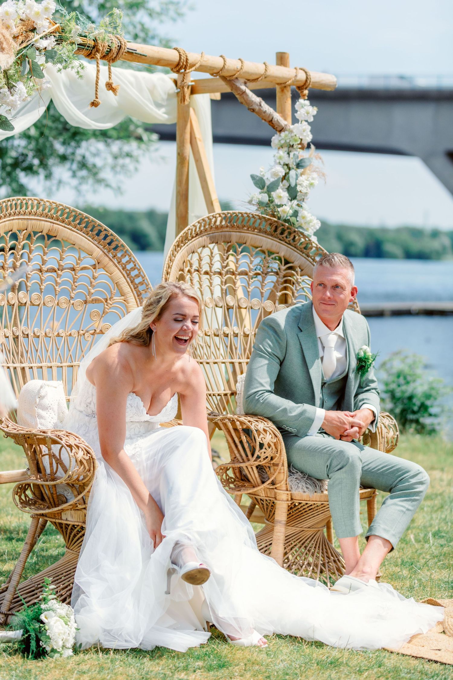Outdoor wedding setup with rattan chairs and floral arch by a waterfront setting.
