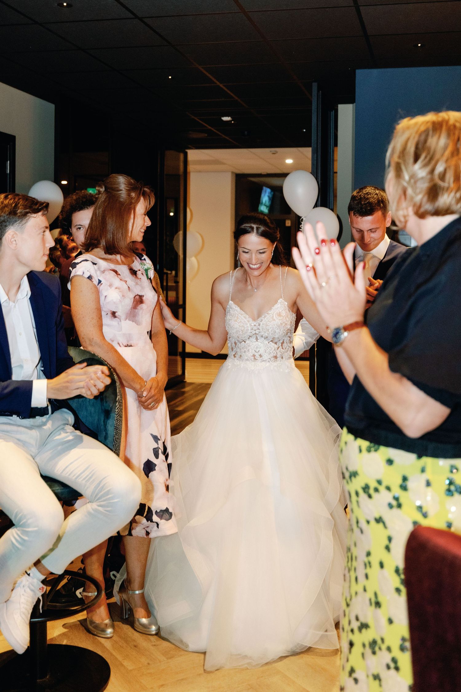 A bride in a flowing white gown laughs joyfully while surrounded by wedding guests at an indoor reception.