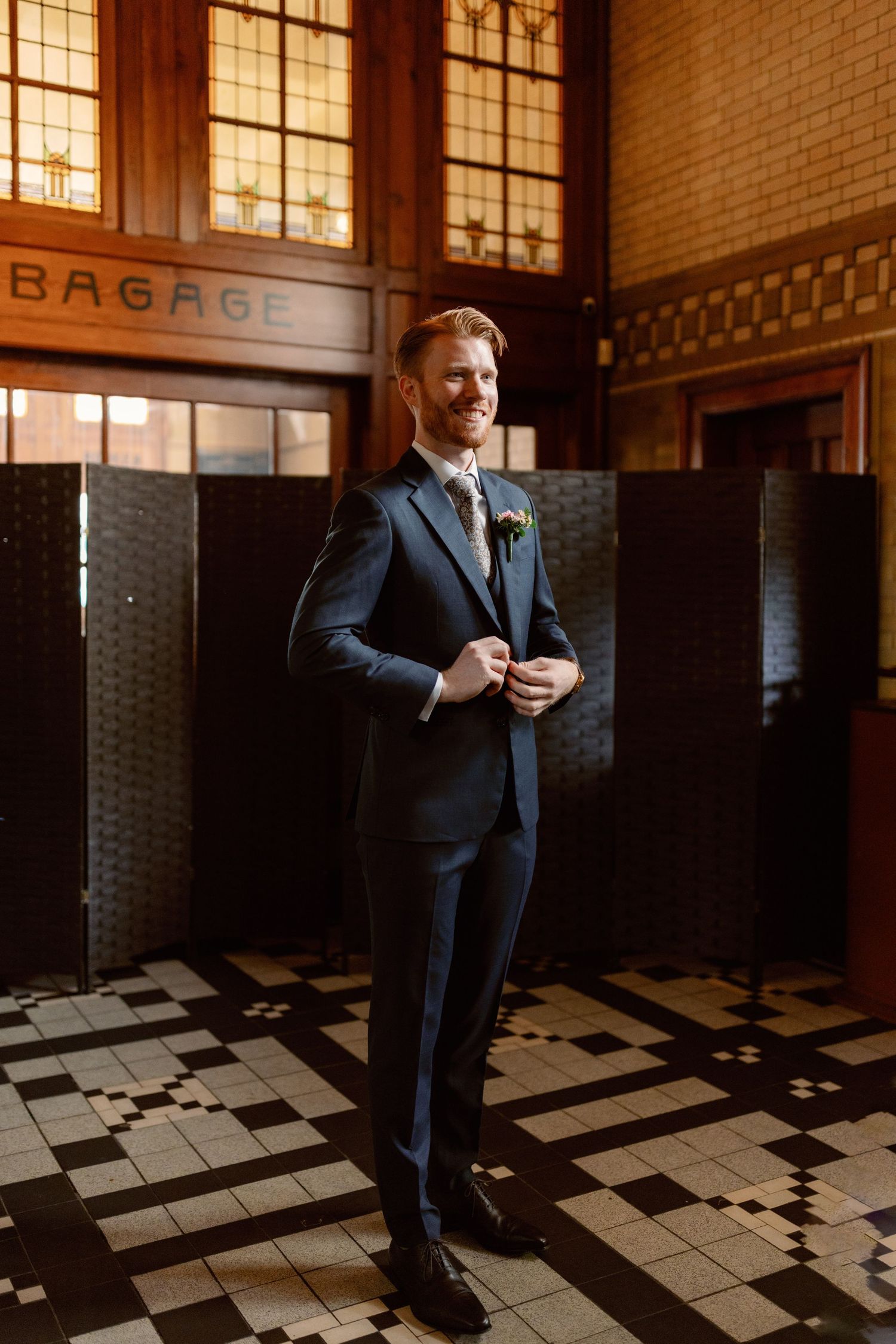 A groom in a dark suit stands in an art deco train station with checkered floors and vintage architectural details.
