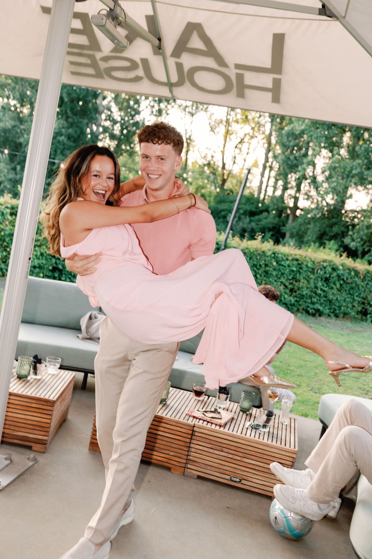 A couple in white and pink outfits dance playfully in a covered outdoor patio space.