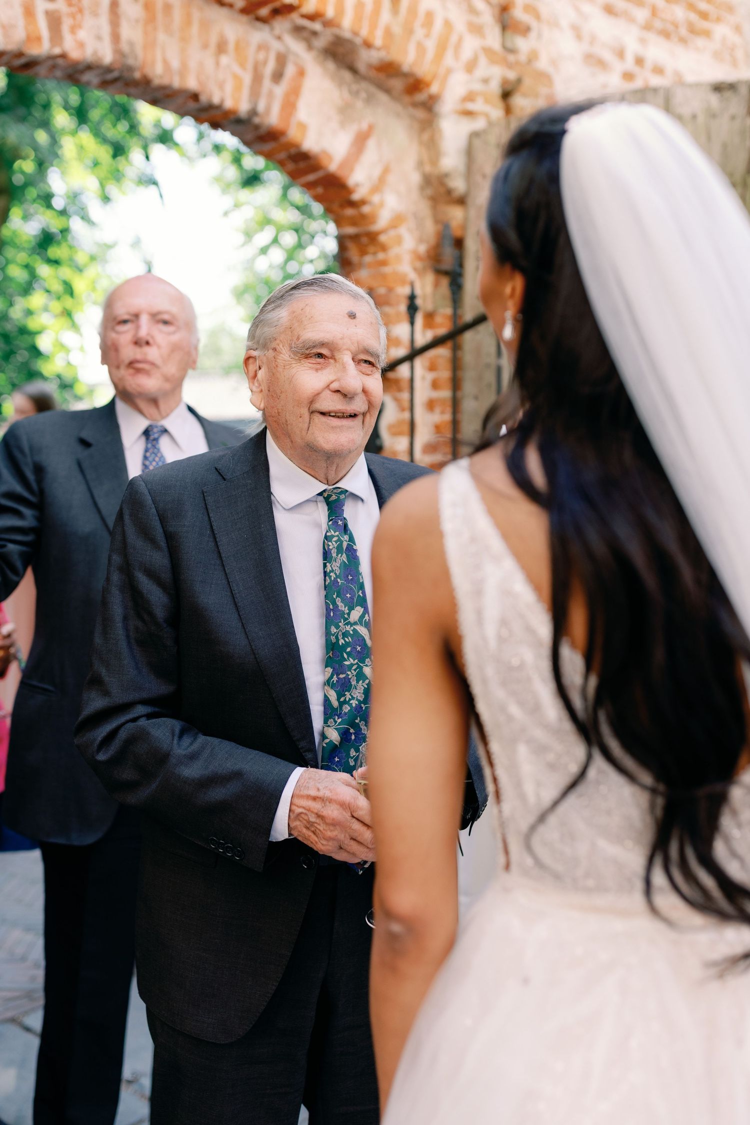 A tender moment at an outdoor wedding ceremony under a brick archway with guests looking on.