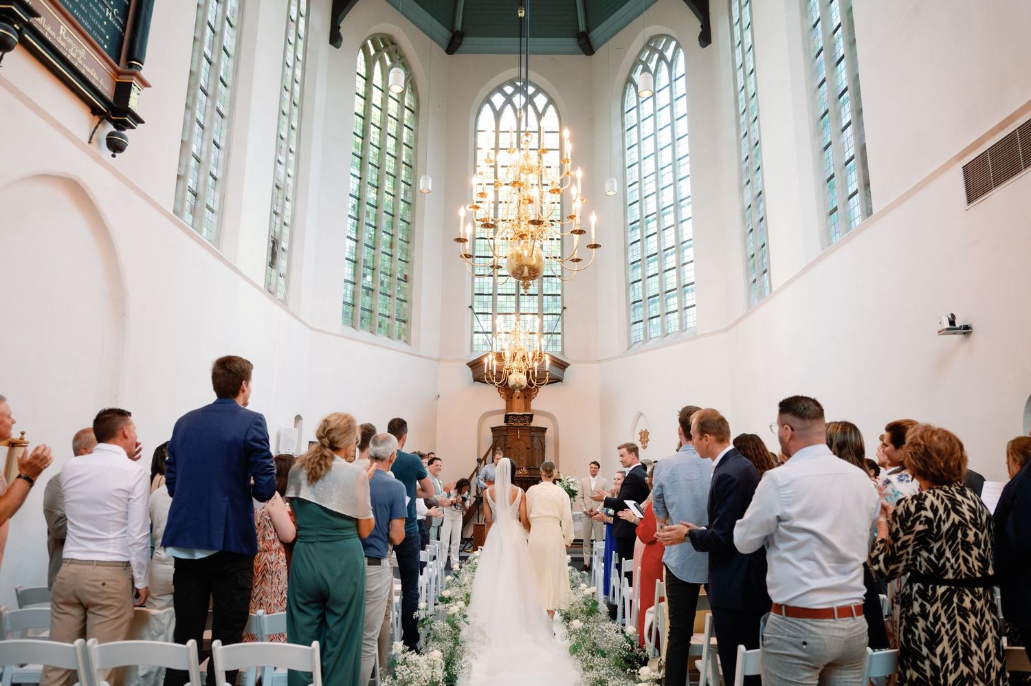 Wedding guests stand in a bright white church interior with stained glass windows during a ceremony.