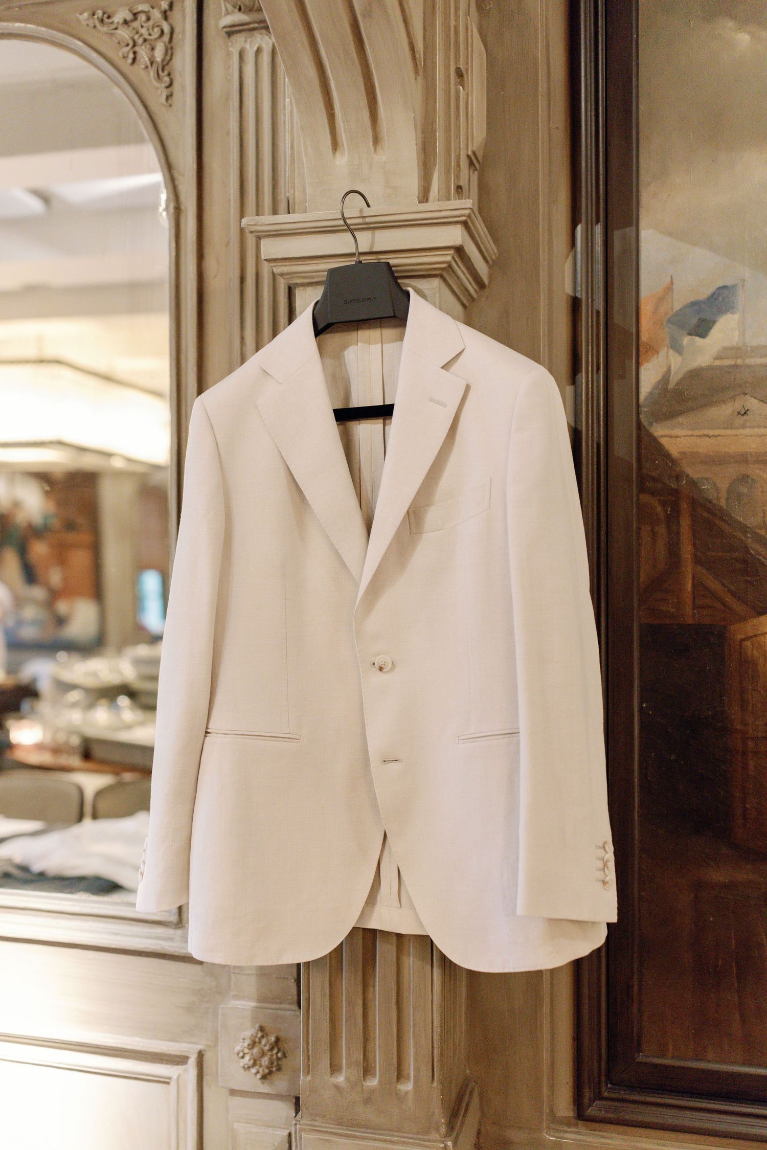 A crisp white formal blazer hanging on a wooden hanger against an ornate architectural backdrop in warm lighting.