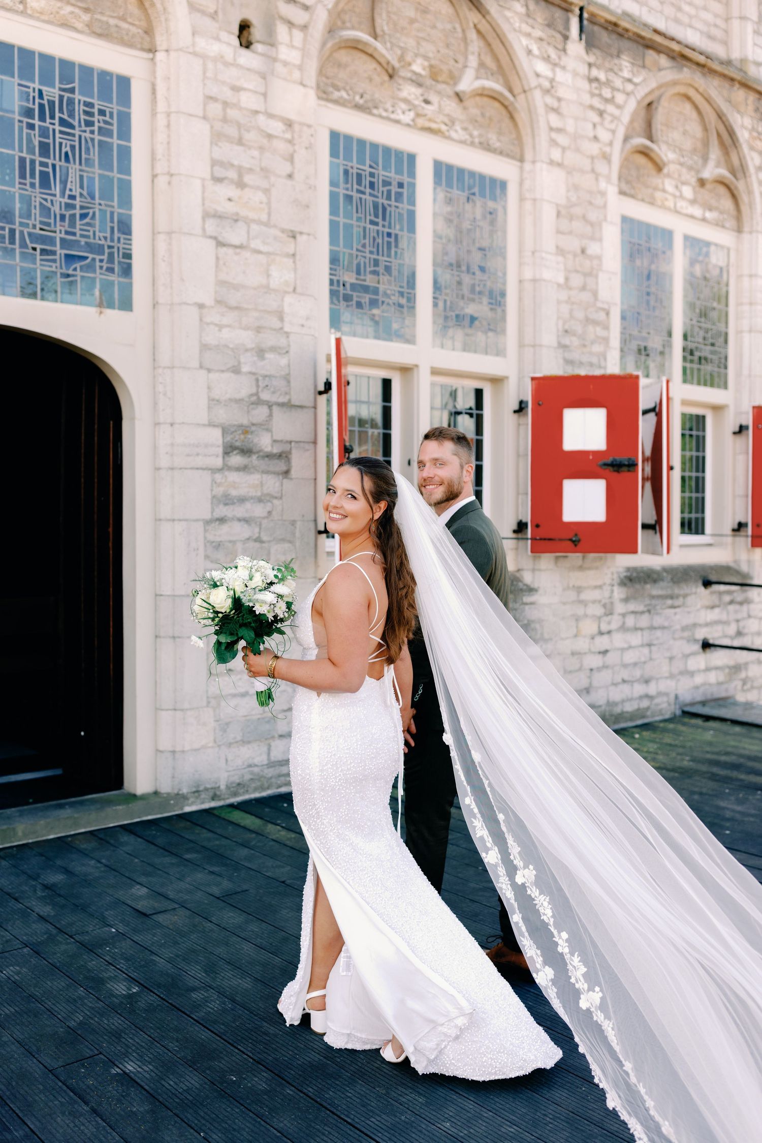 A couple in wedding attire share an intimate moment against a stone building with blue windows and a flowing white veil.