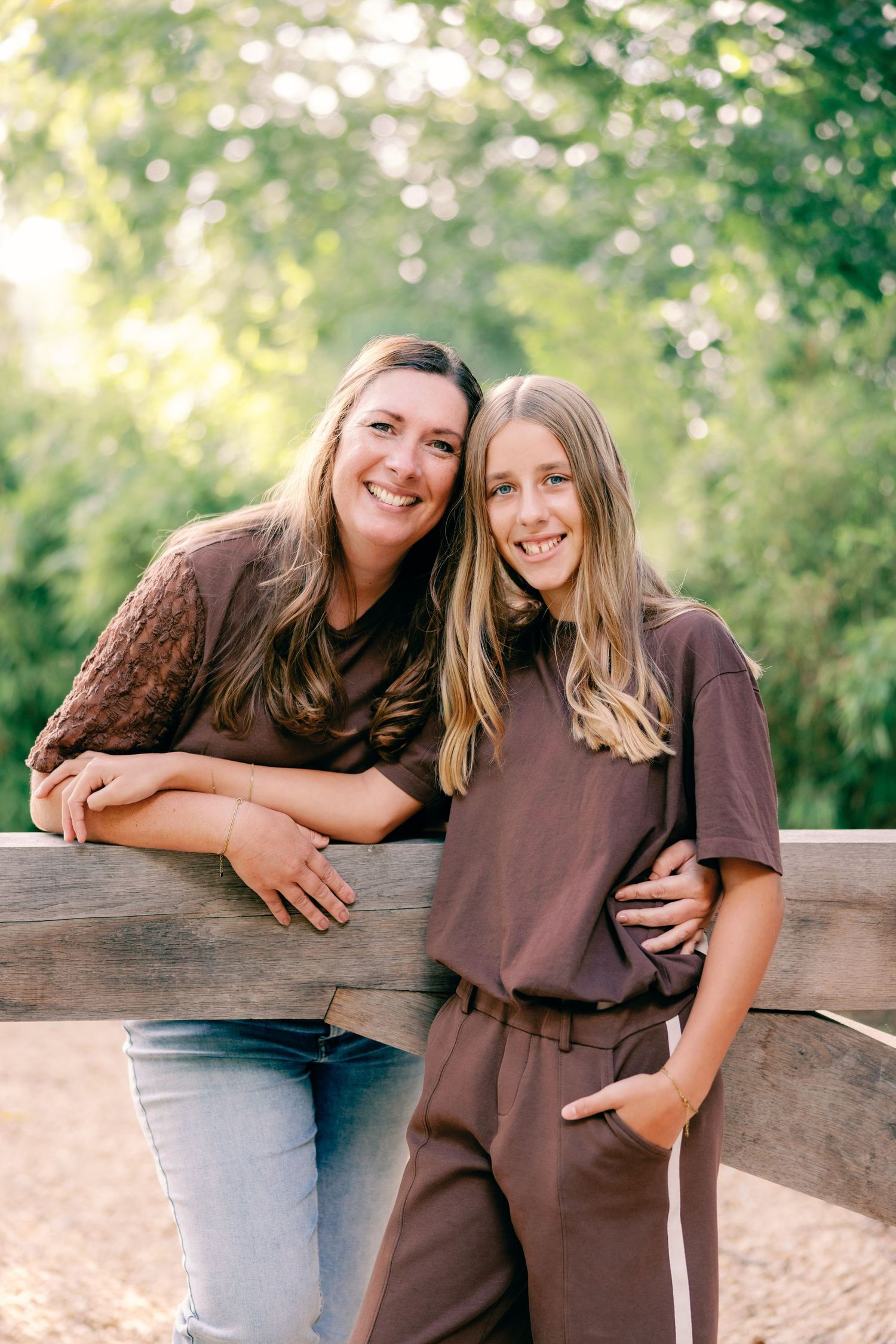 Smiling woman and young girl with long hair in matching brown outfits posing together against a rustic wooden fence in a sunny park.