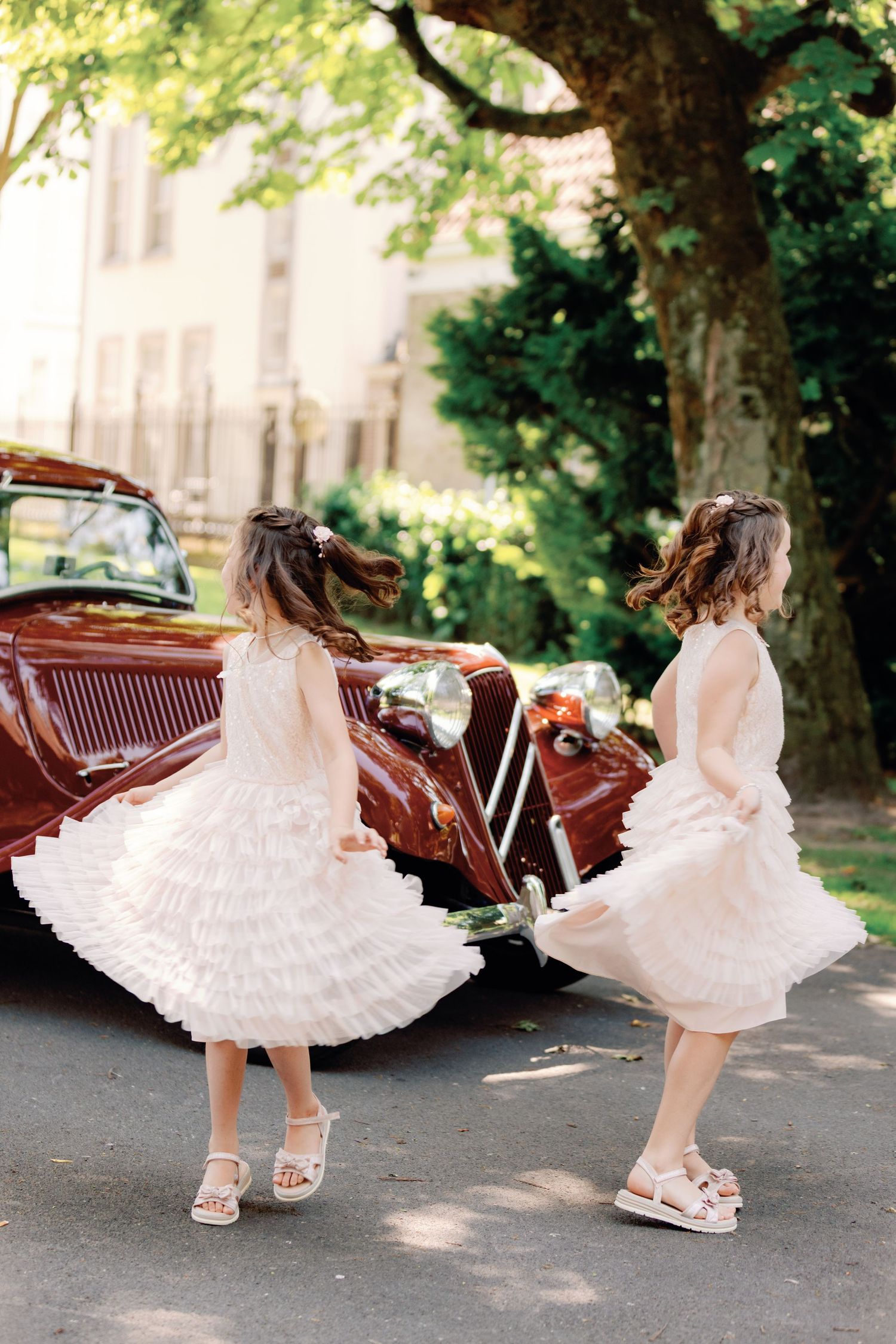Two girls in white dresses twirl near a vintage red car outdoors.