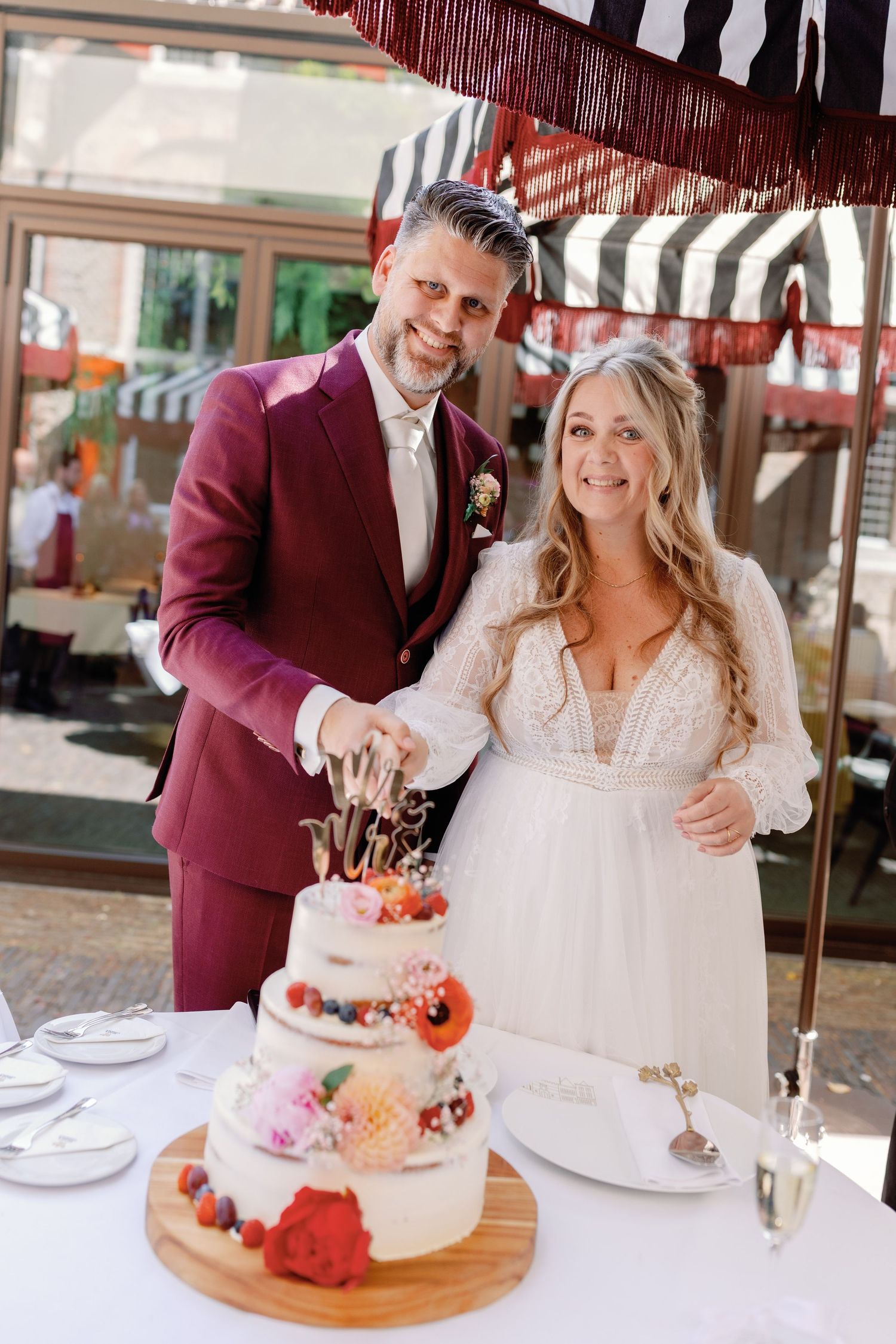 A wedding couple cuts into their three-tiered white cake decorated with pink and orange flowers under a striped awning.