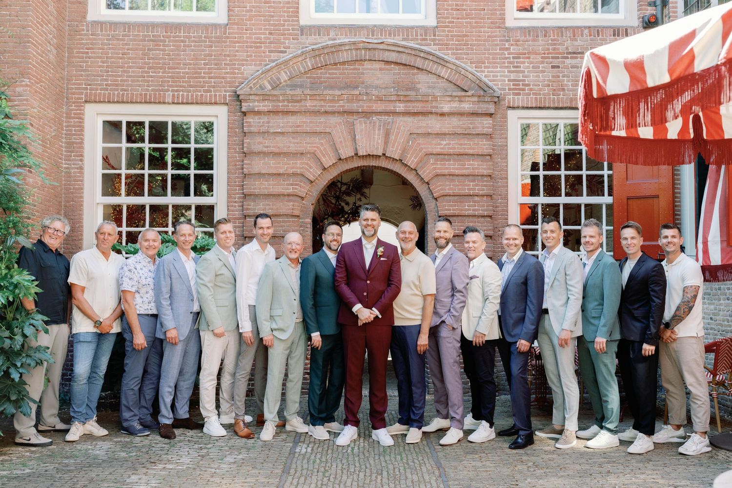 A group of groomsmen in coordinated suits and sneakers pose together outside a brick building entrance.