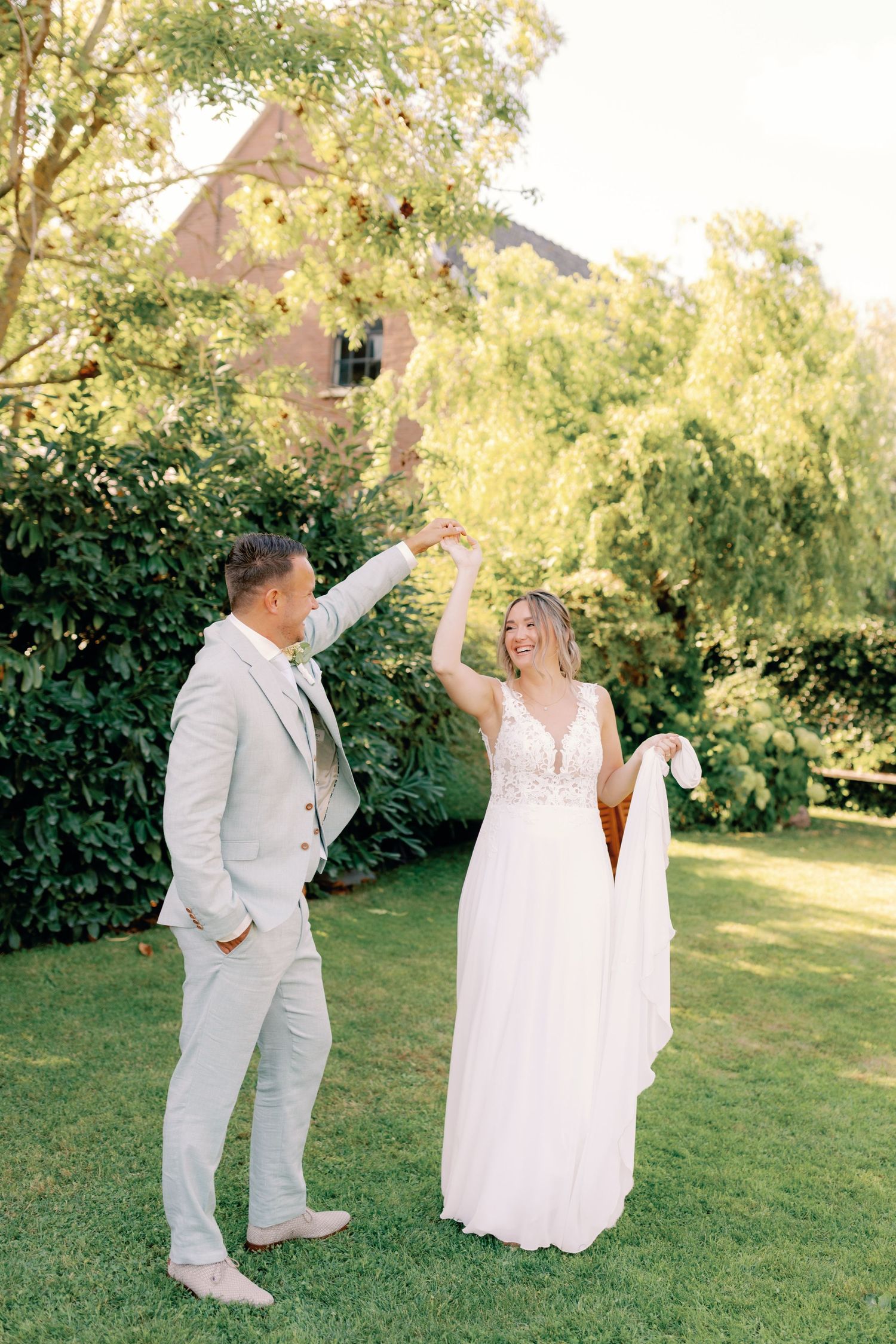 A couple in white wedding attire playfully dancing together on a green lawn with trees in the background.