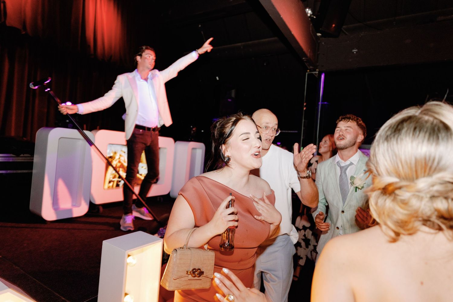 A lively wedding reception dance floor with guests dancing and celebrating under red mood lighting.