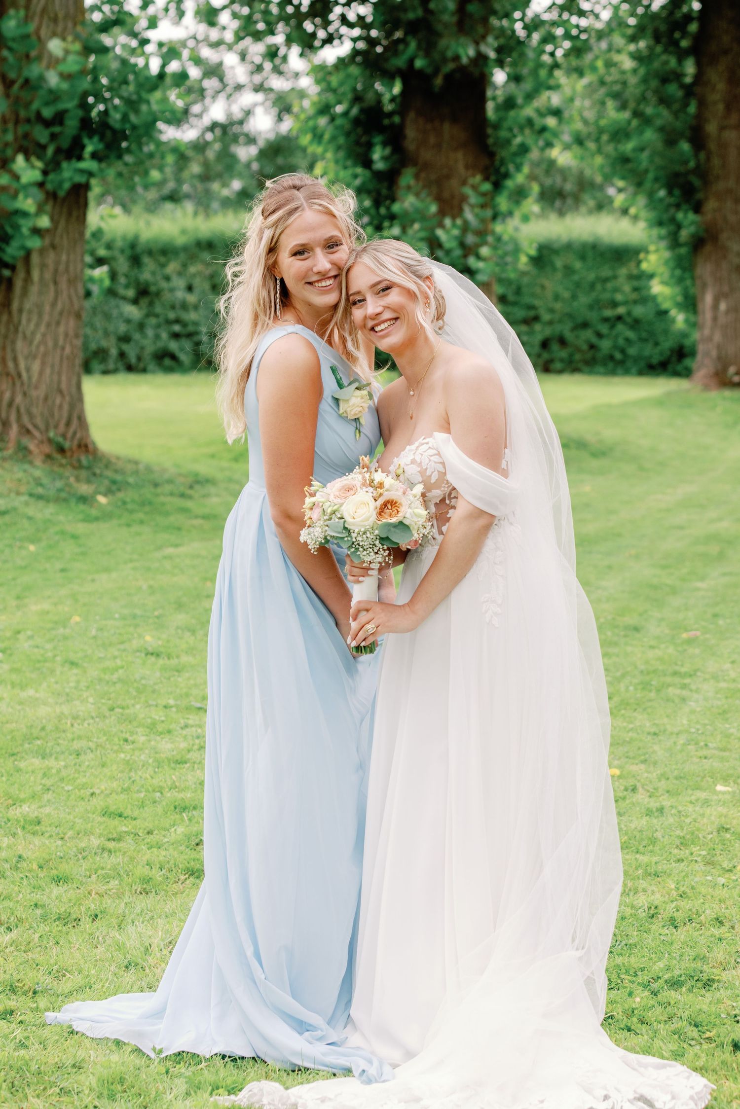 A bride in white and bridesmaid in light blue share a joyful moment together on a lush green lawn.