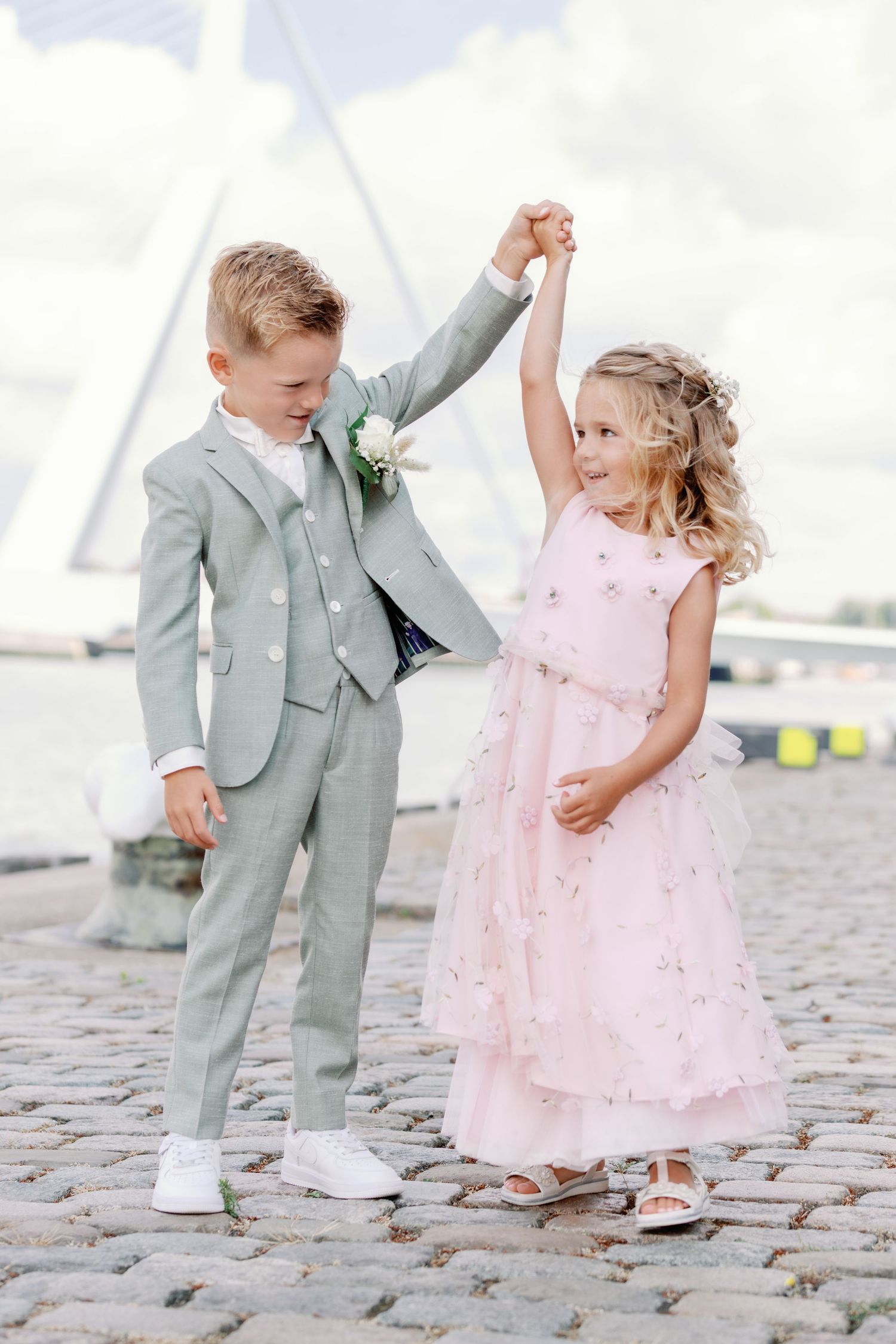 Two children in formal attire dance and twirl together on a cobblestone walkway wearing pink dress and gray suit.