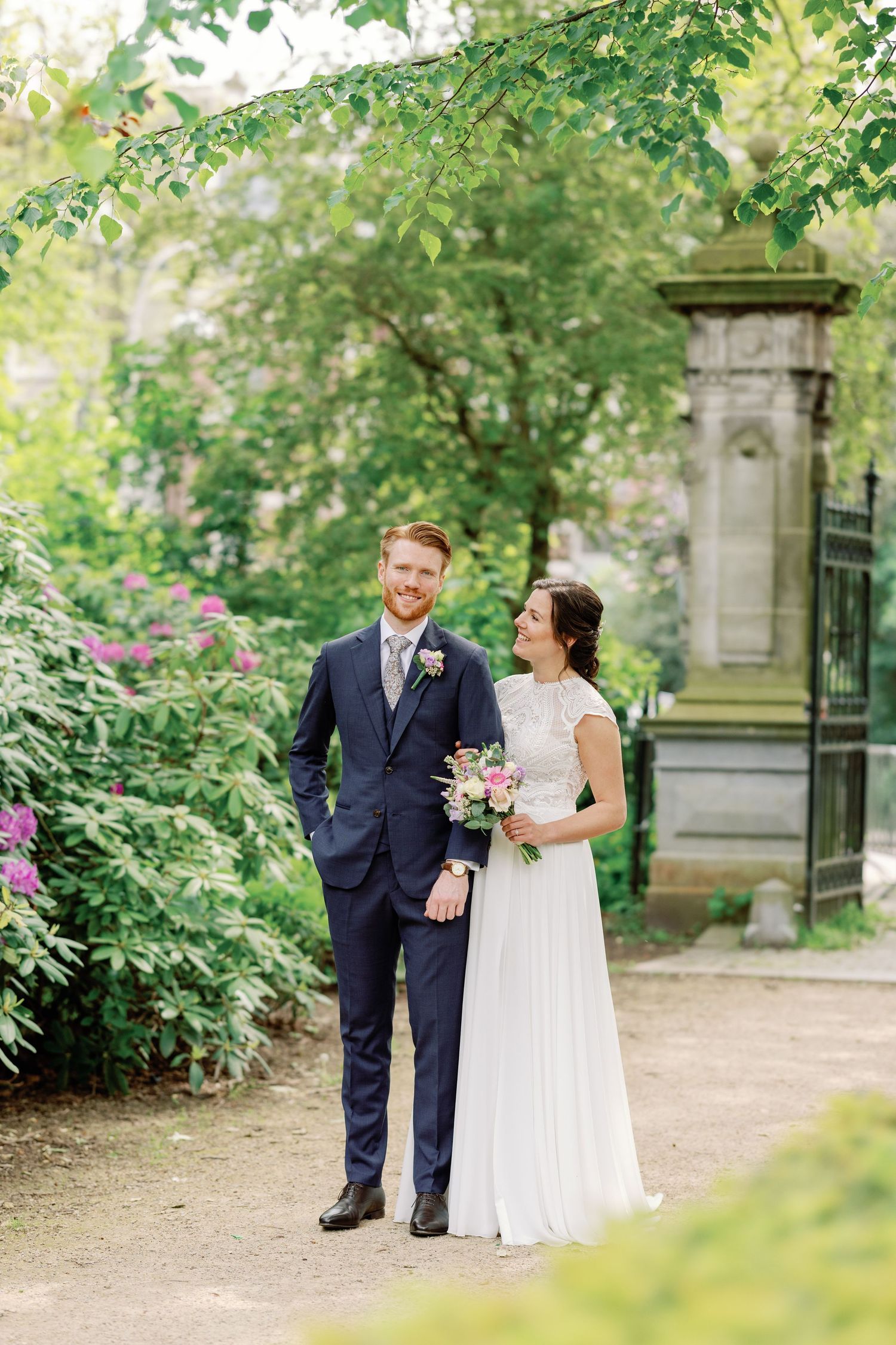 A couple in wedding attire walks along a garden path surrounded by lush greenery on a bright summer day.
