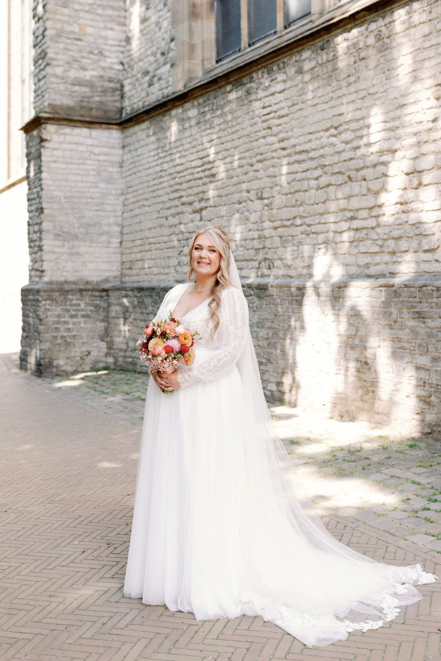 A bride in a flowing white lace gown with long veil holds a peach and coral bouquet against a rustic brick wall.