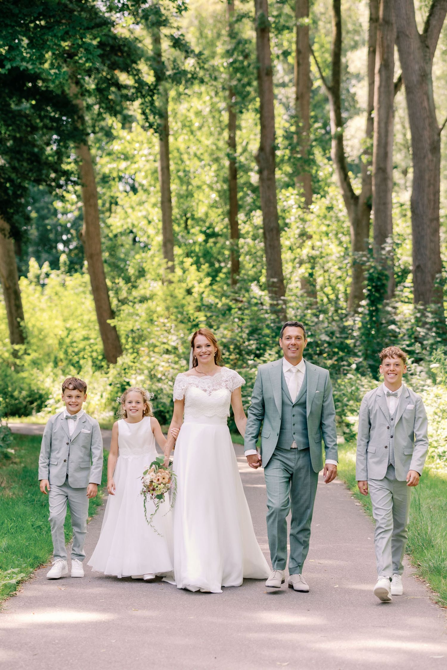 A wedding party in gray suits and white dresses walks along a tree-lined path in a sunlit garden setting.
