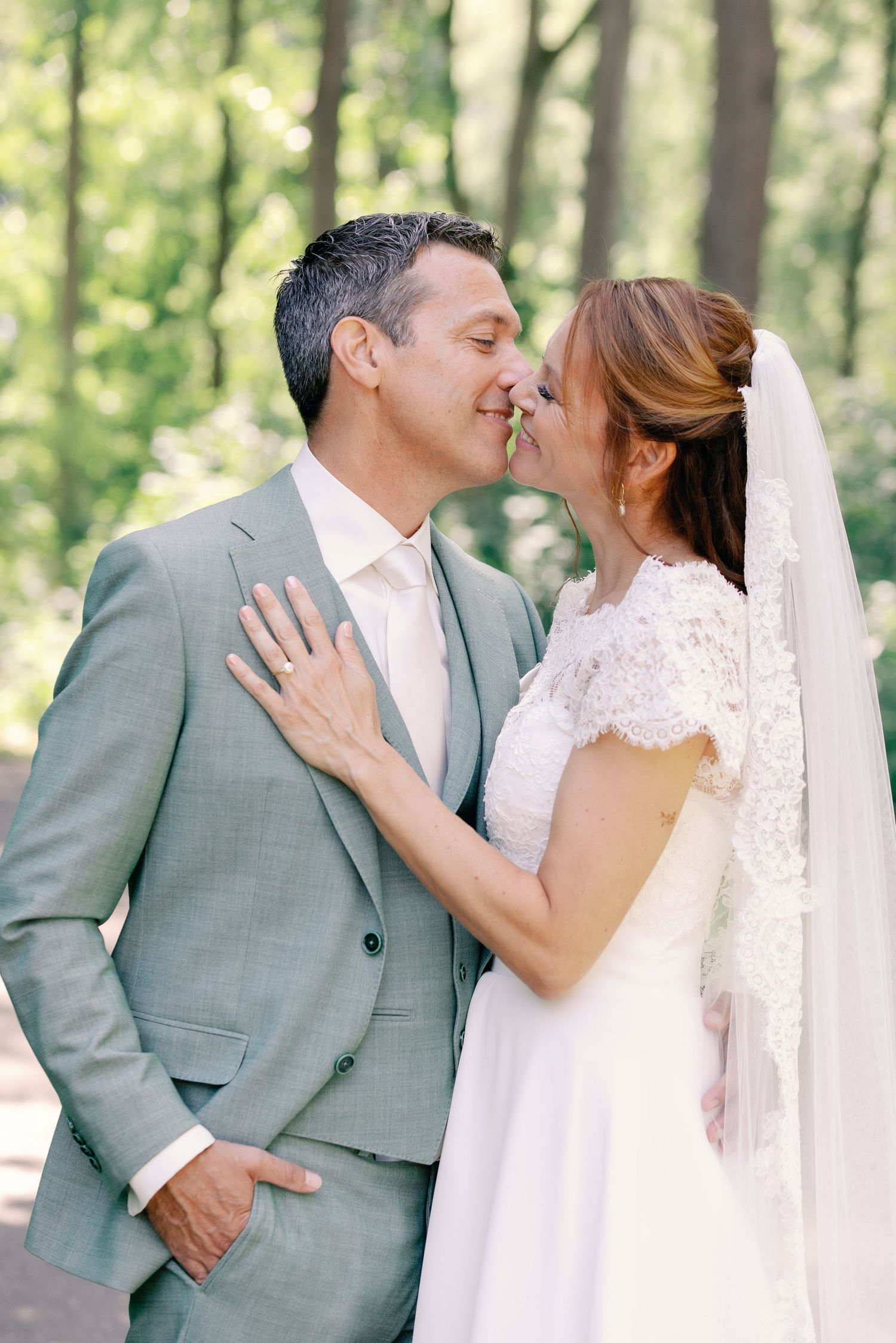 A romantic wedding couple shares intimate kisses in a sunlit forest setting wearing gray suit and lace dress.