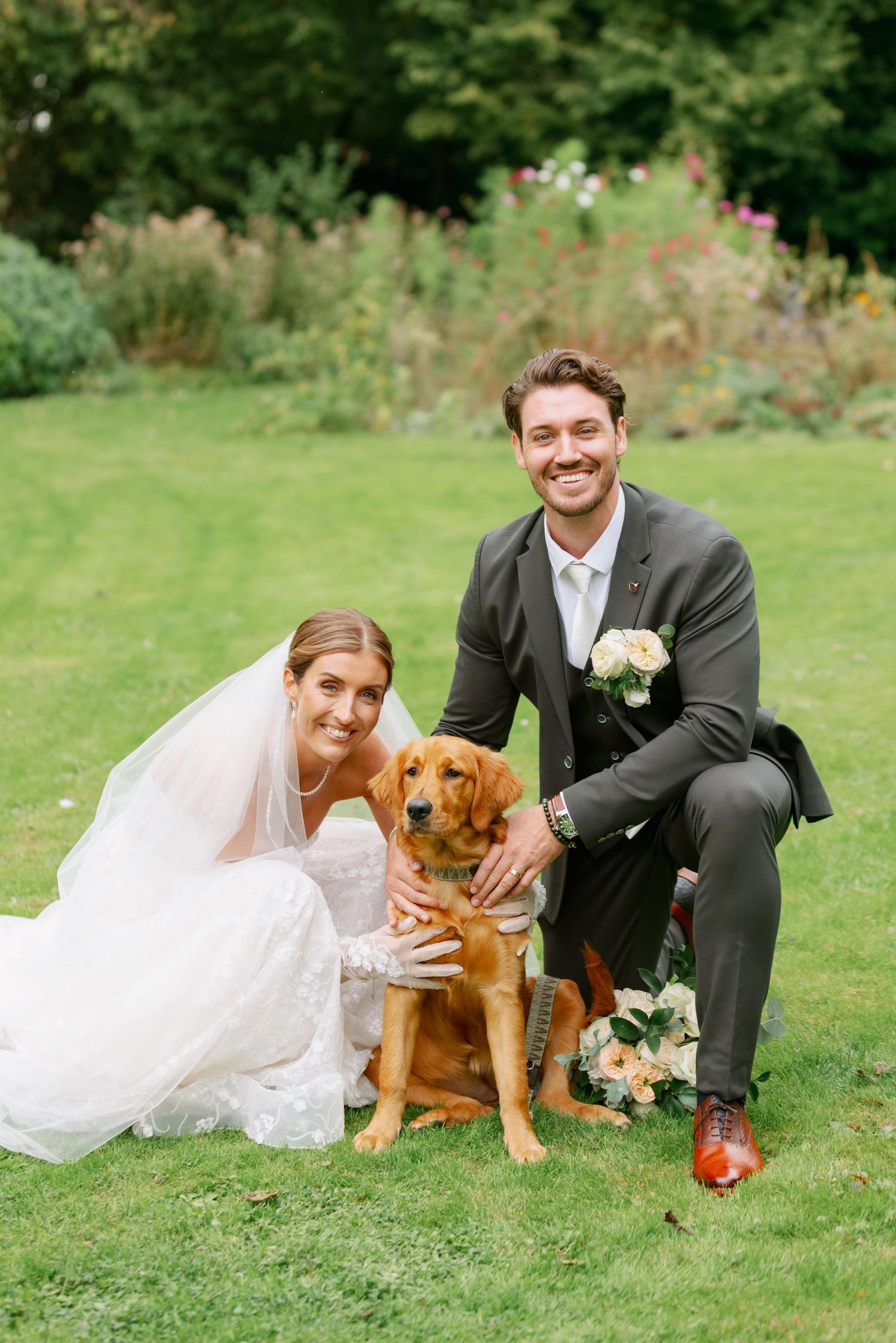 A couple in wedding attire poses playfully on grass with their golden brown dog during outdoor photo session.