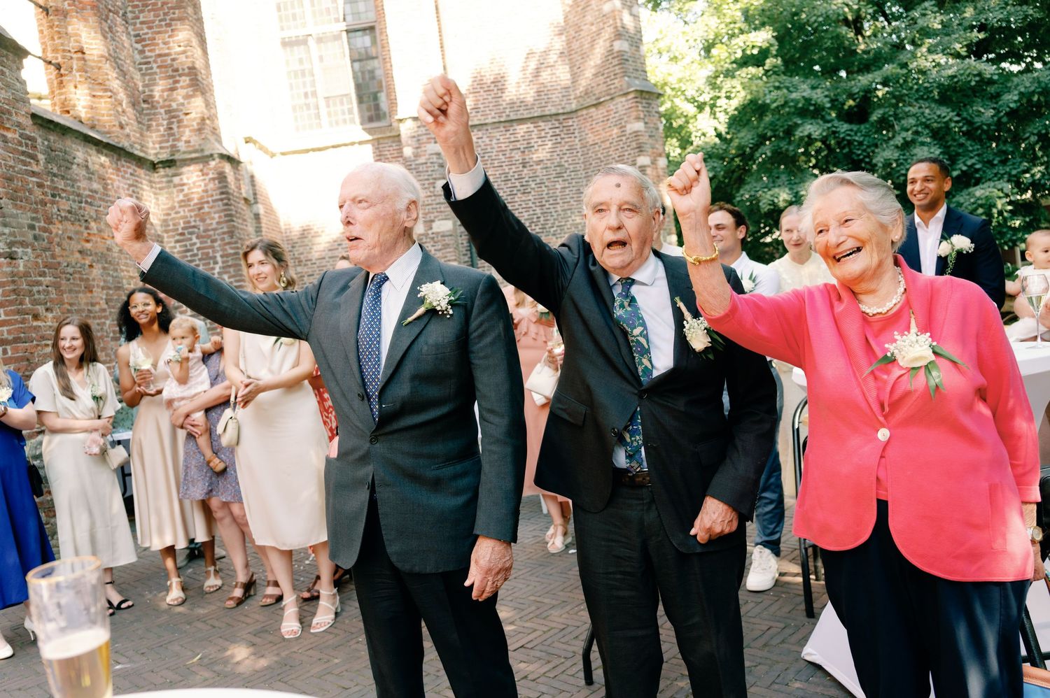 A group of wedding guests in formal attire celebrate joyfully with raised arms outside a historic stone church.