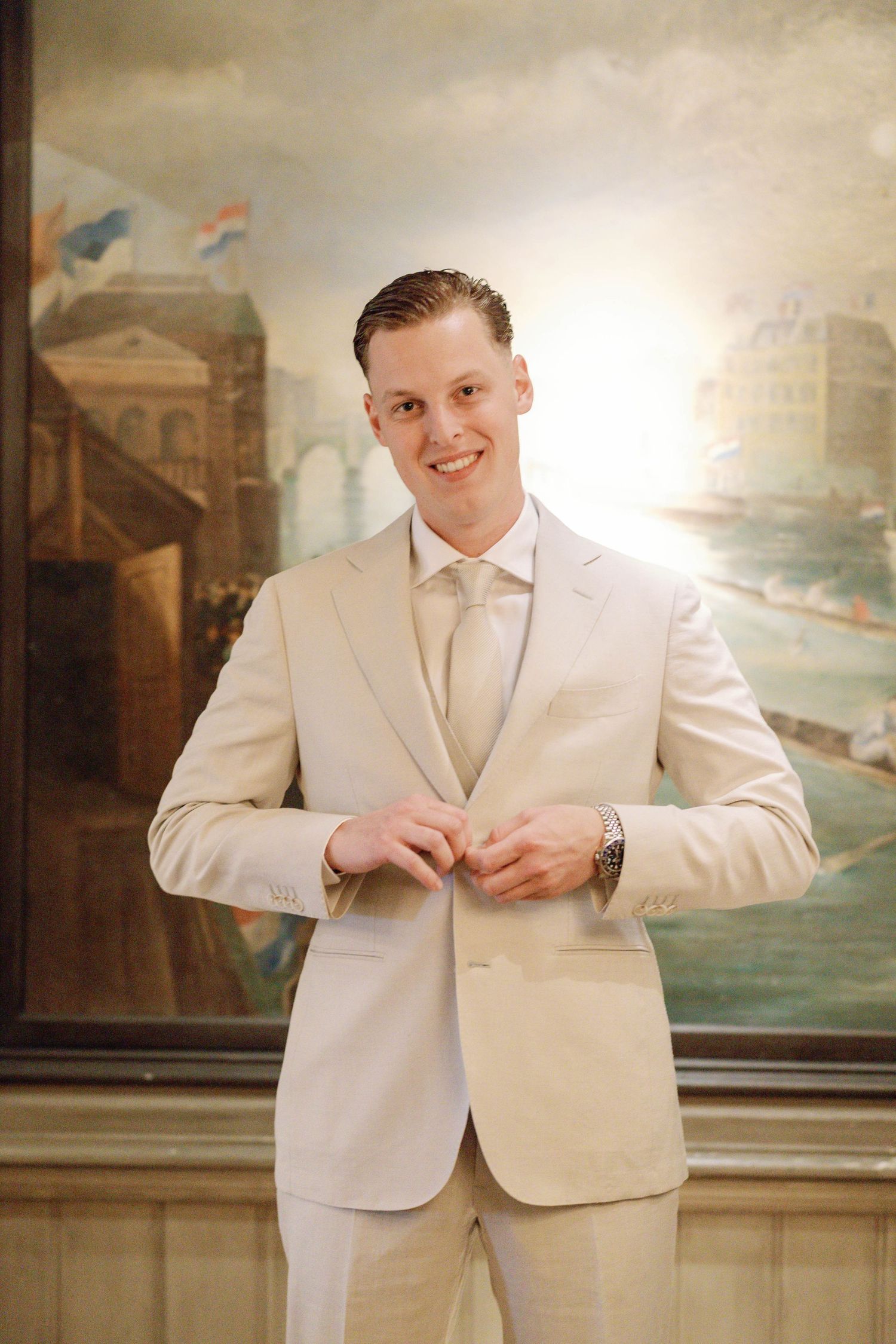A groom in a cream-colored suit and tie adjusts his jacket while standing in front of a scenic painted backdrop.