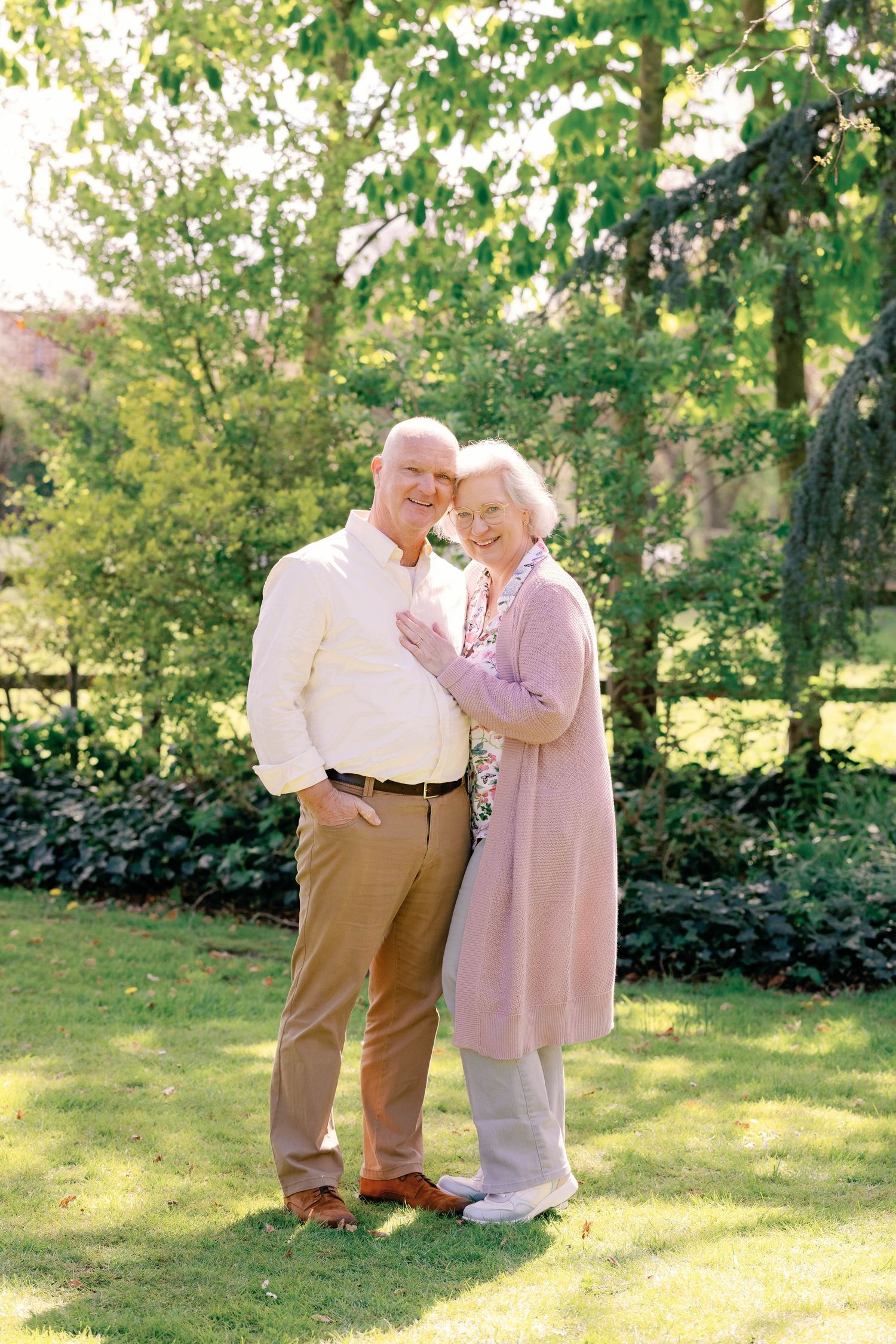 Elderly couple smiling and embracing in a lush green garden on a sunny day.