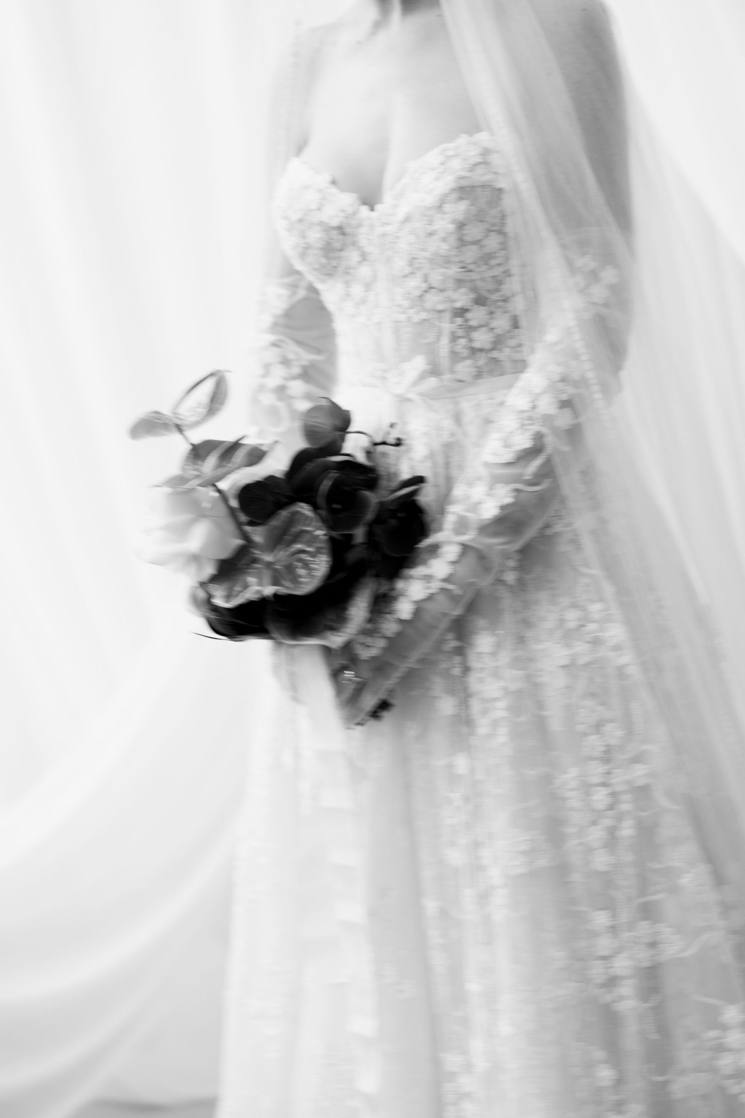 Black and white photo of a bride in lace gown holding a floral bouquet with roses, draped in a flowing veil.