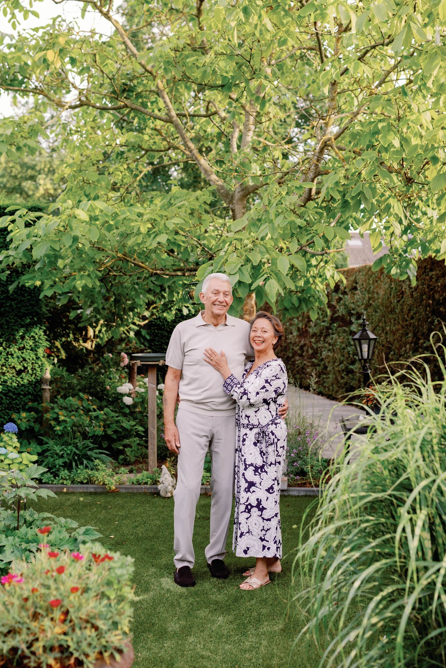 Elderly Asian couple smiling and embracing in a lush green garden surrounded by colorful flowers and trees.