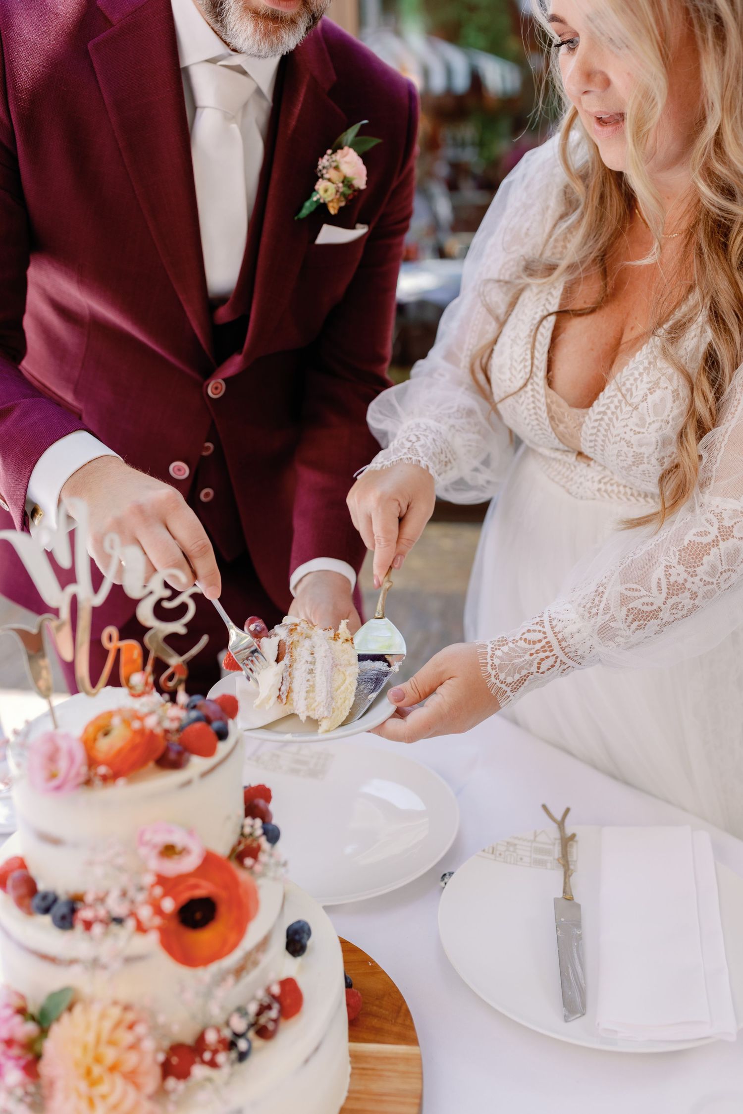 A couple cuts into their elegant white wedding cake decorated with fresh berries and flowers at their burgundy-themed reception.