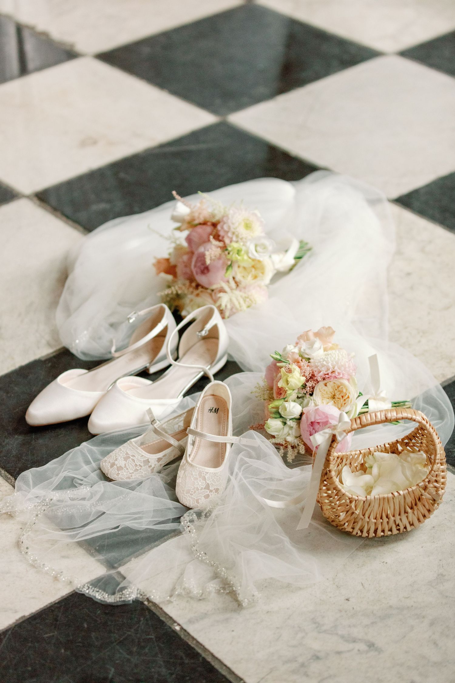 Wedding accessories including white bridal shoes, delicate veil, and floral bouquets arranged on checkered marble floor.