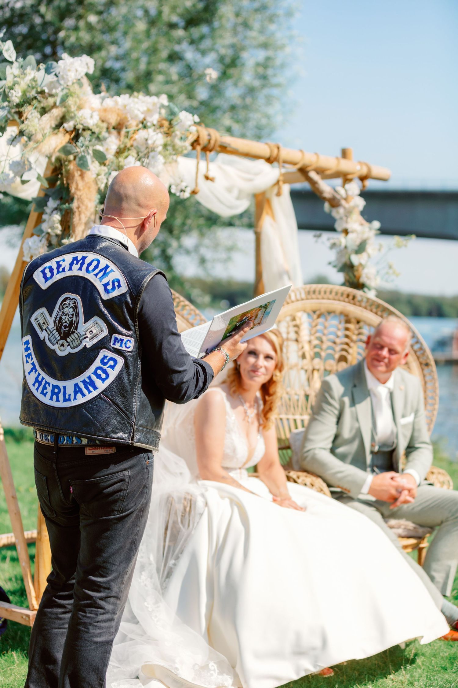 Wedding officiant in leather vest conducts ceremony under floral arch by waterfront.