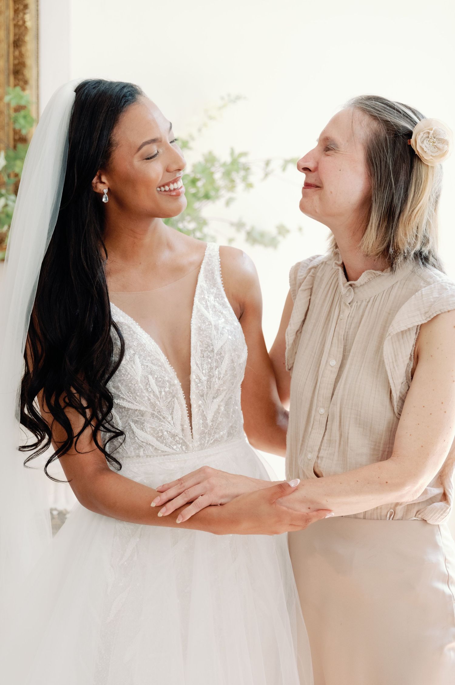 A tender moment shared between a bride in a white lace wedding dress and a companion in beige, both smiling joyfully.