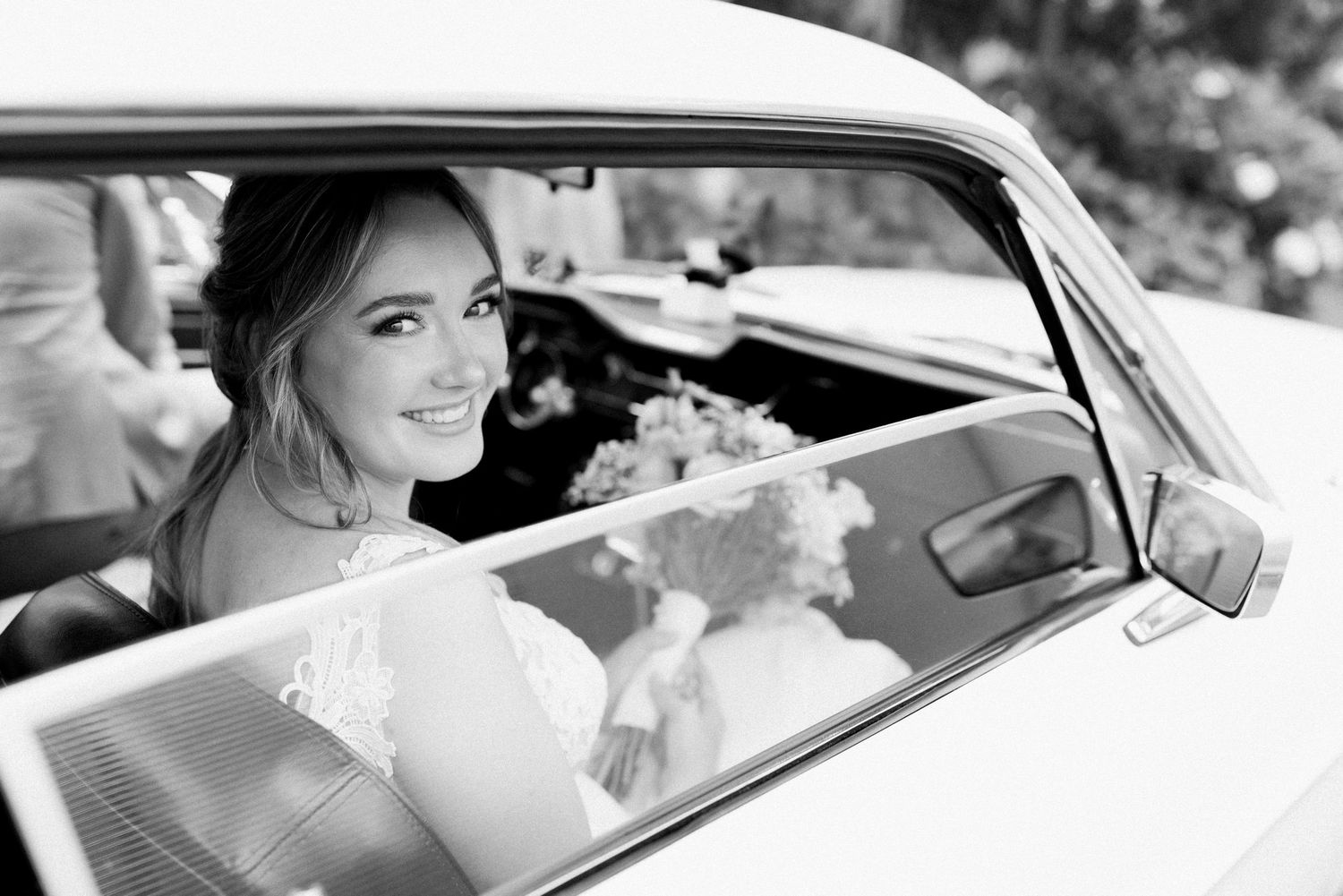 Black and white photo of a bride smiling from inside a classic car on her wedding day.