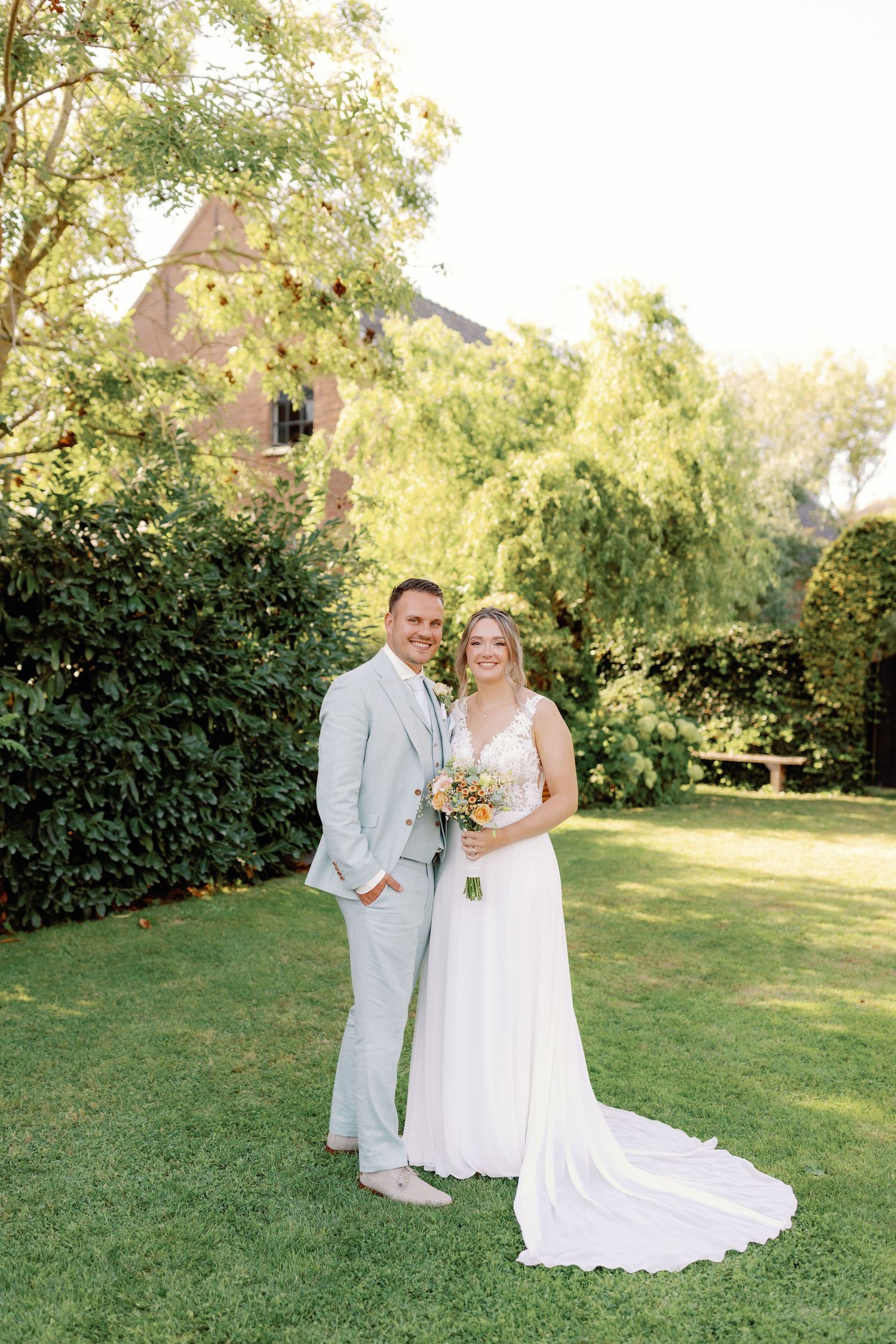 A couple in wedding attire poses together on a sunny lawn with green hedges in the background.