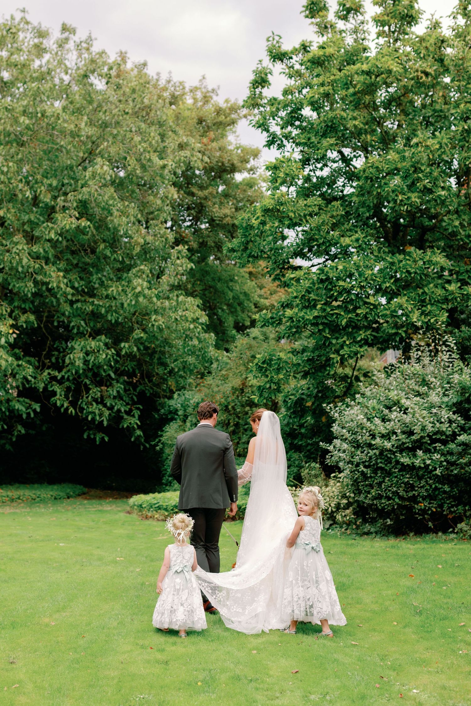 A wedding party walks away from the camera on grass with two small children holding the bride's train.