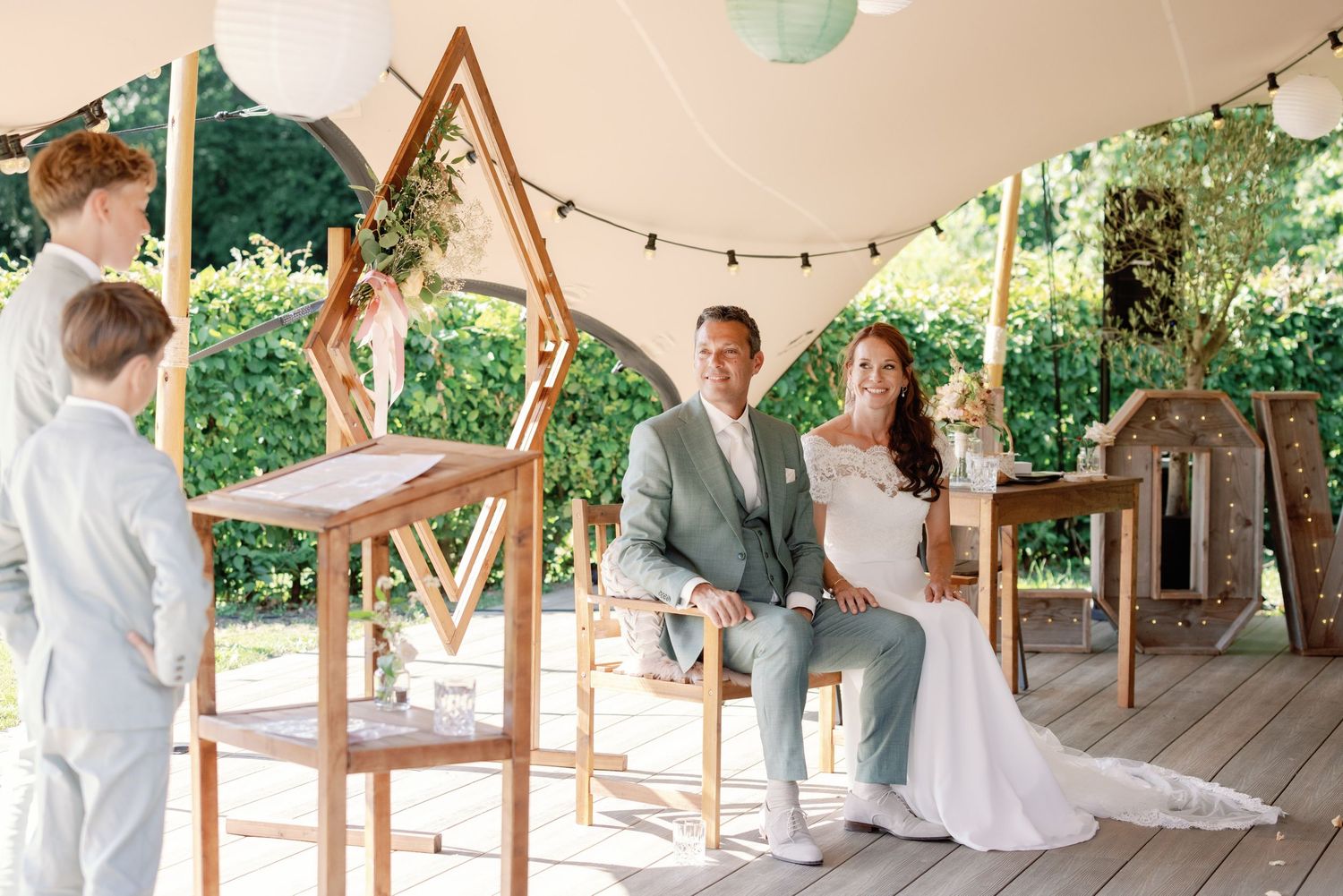 A couple sits under an outdoor tent canopy during a rustic garden wedding ceremony with wooden decor.