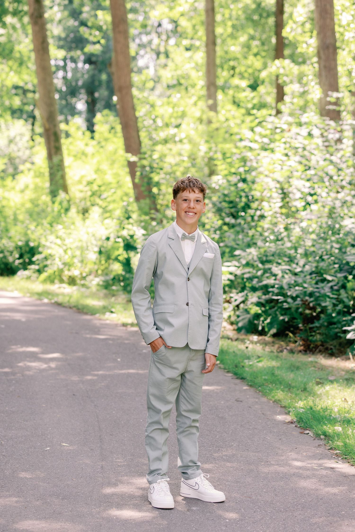 A person in a light gray suit and white sneakers stands on a sunlit forest path surrounded by lush greenery.