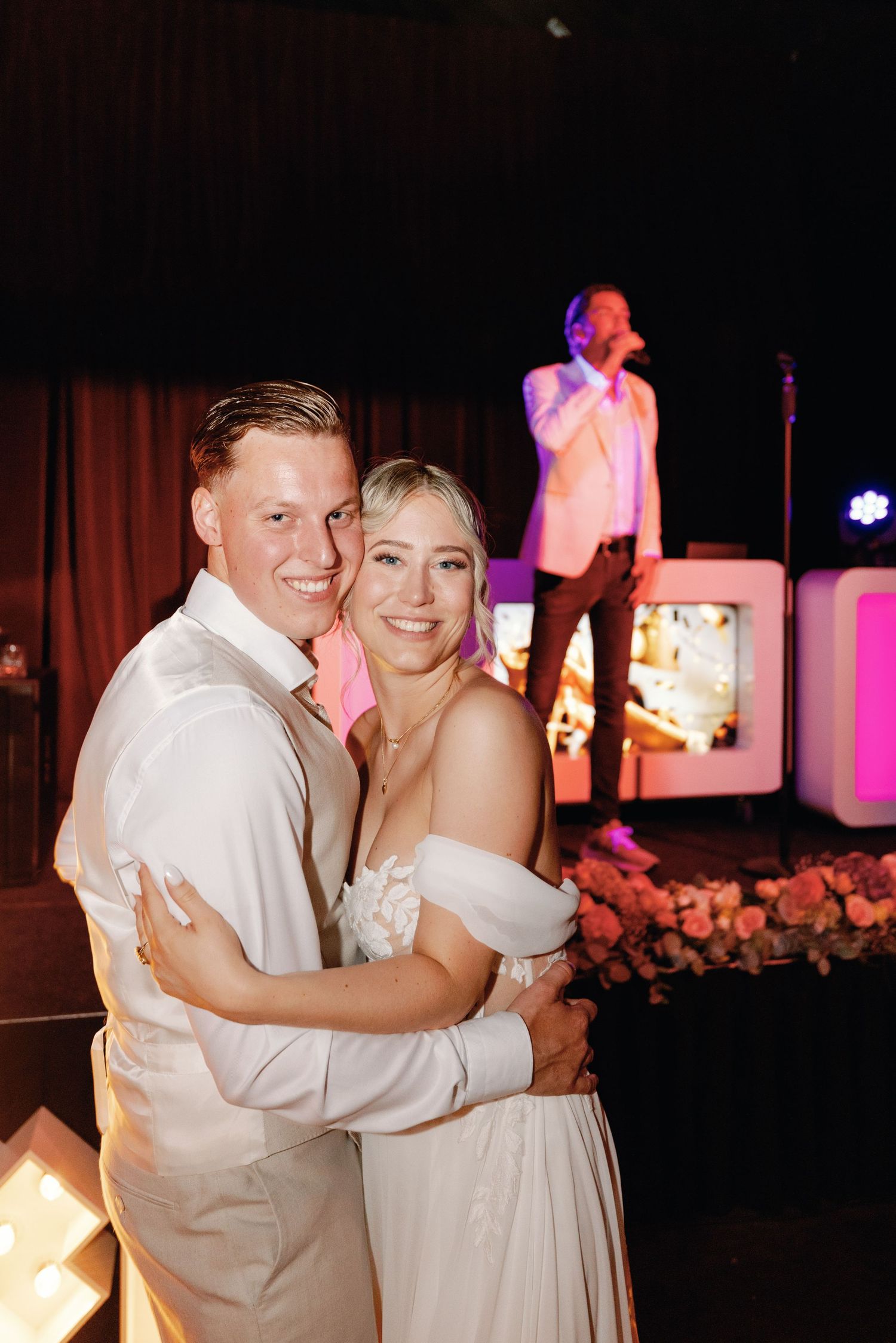 A couple shares a romantic dance together at an evening reception with pink uplighting and white decor in the background.
