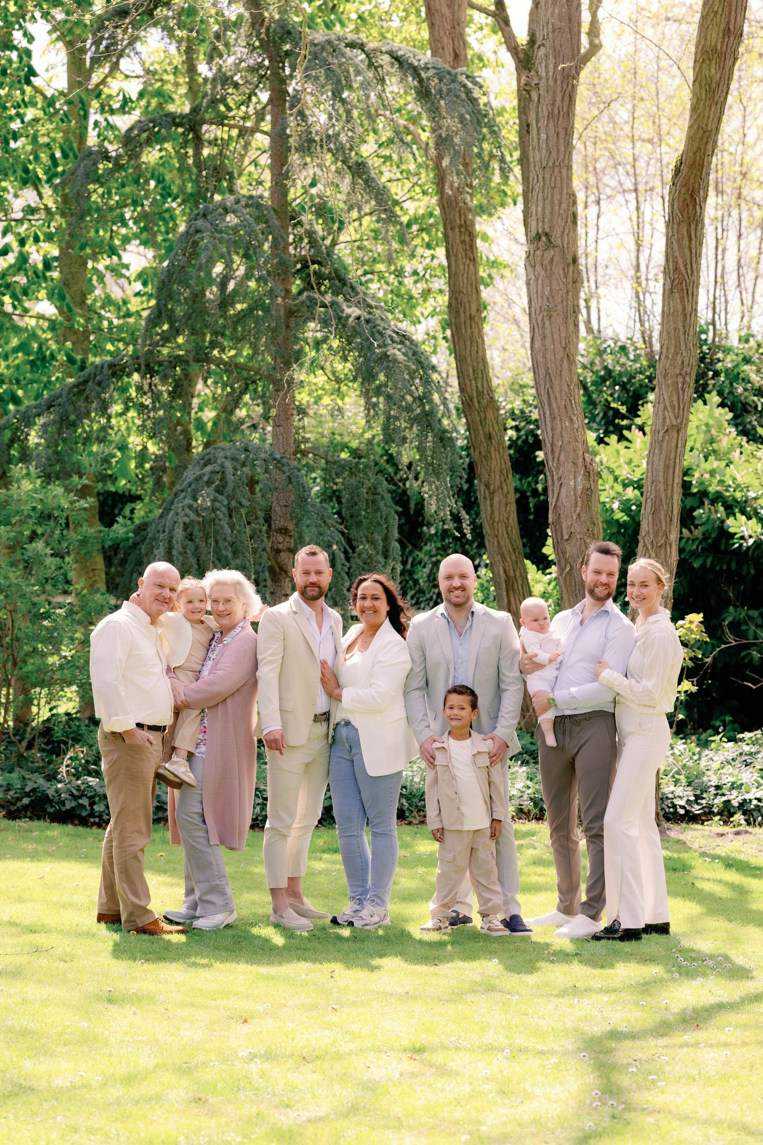 Multi-generational family posing together outdoors in a lush green garden, dressed in elegant light-colored outfits.
